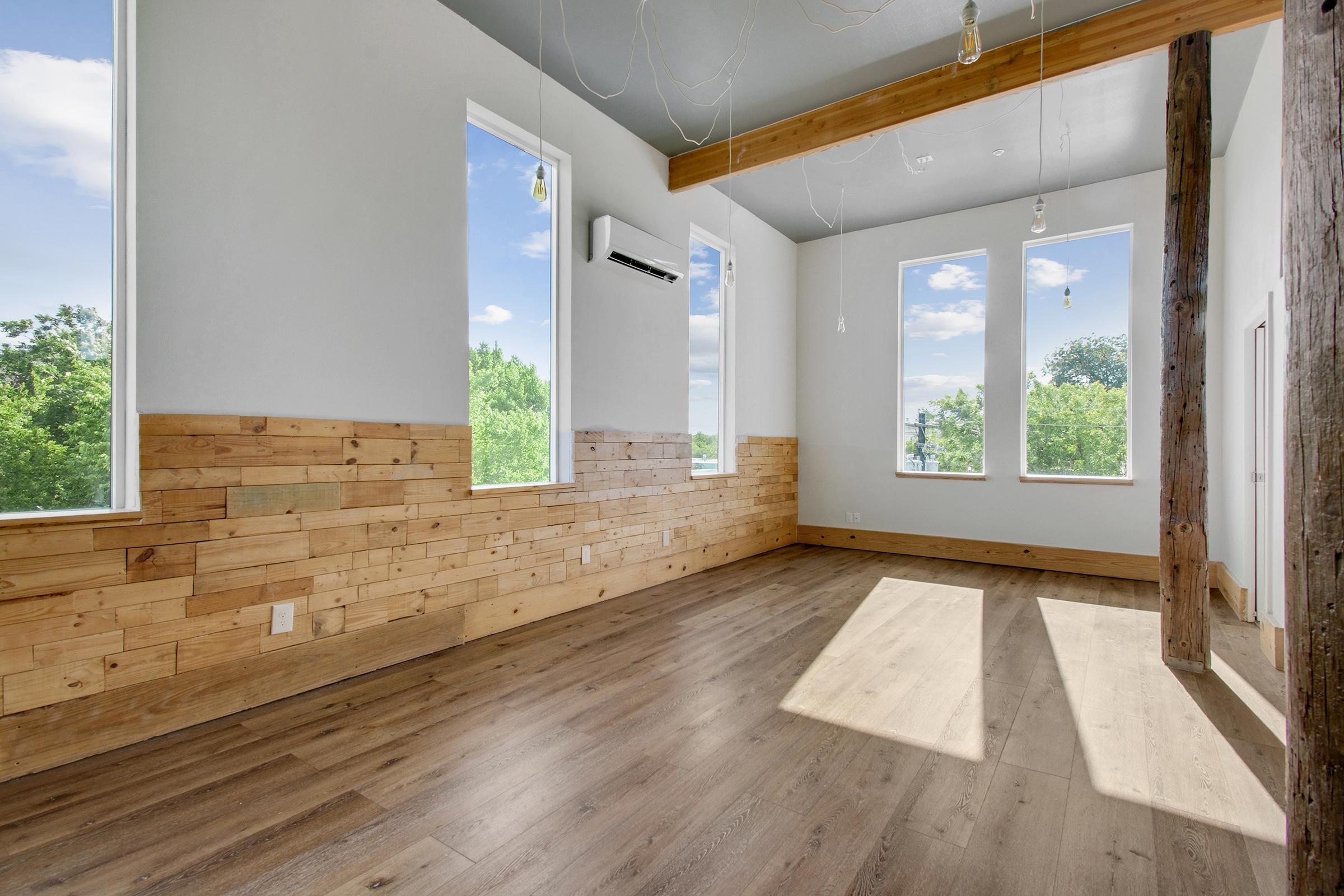 Bright, modern interior of a room featuring large windows with scenic views. The walls have a mix of wood paneling and white paint, and there are wooden beams and hanging light fixtures. The floor is finished with light-colored wood, creating a spacious and airy atmosphere.