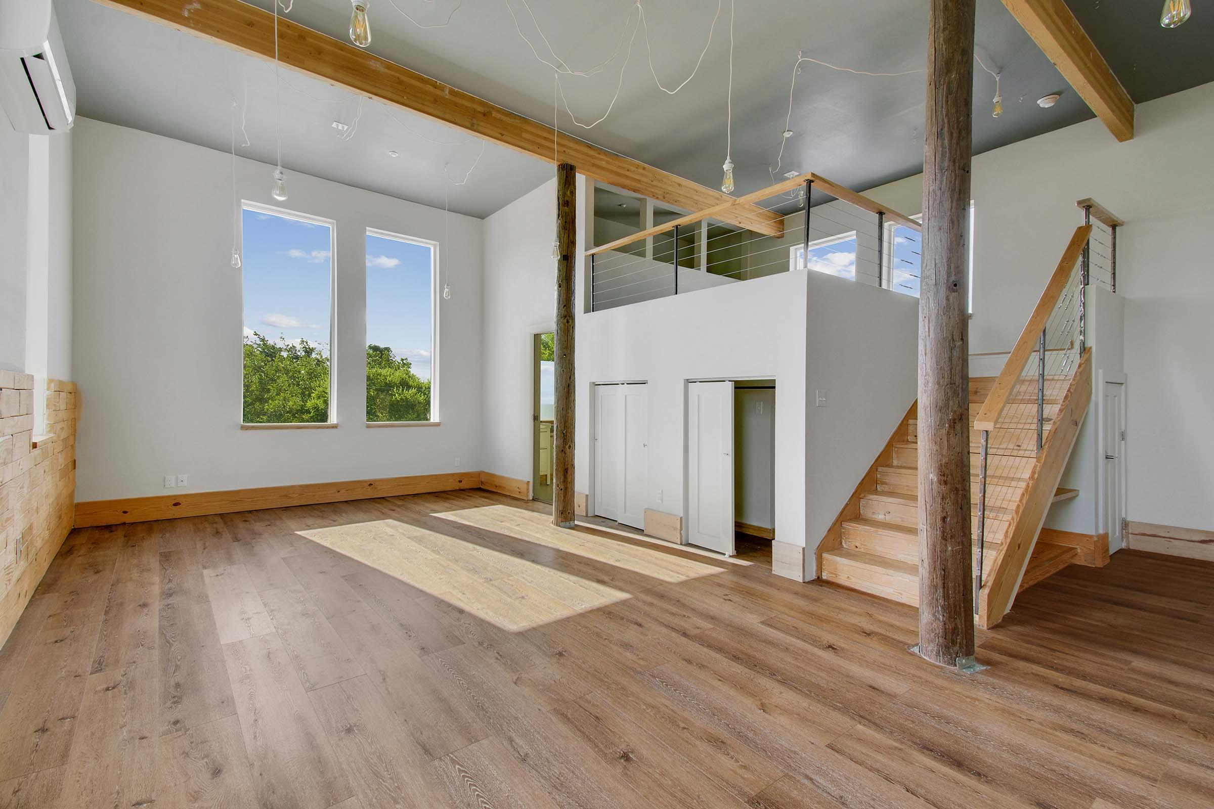 A spacious, modern interior of a house featuring large windows that allow natural light, light wooden flooring, a staircase leading to an upper level, and minimalist decor. Exposed wooden beams and several hanging light bulbs add character to the room.