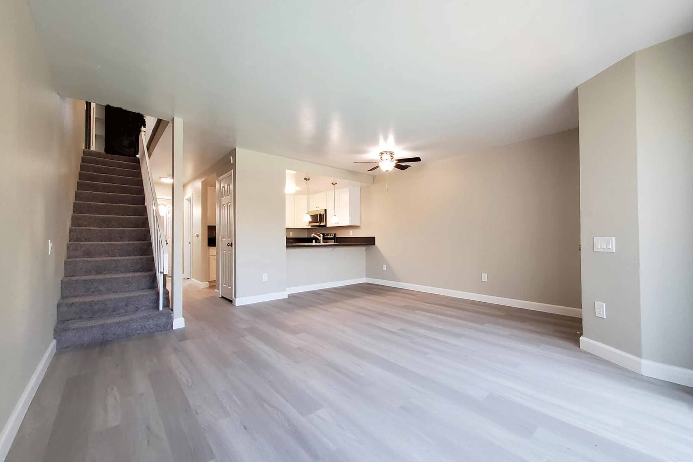 Spacious, modern living room featuring light gray walls, laminate flooring, and a ceiling fan. A staircase is visible on the left, leading to the upper level. The kitchen is open and connected to the living area, with white cabinetry and appliances. Natural light fills the space, creating a bright and inviting atmosphere.