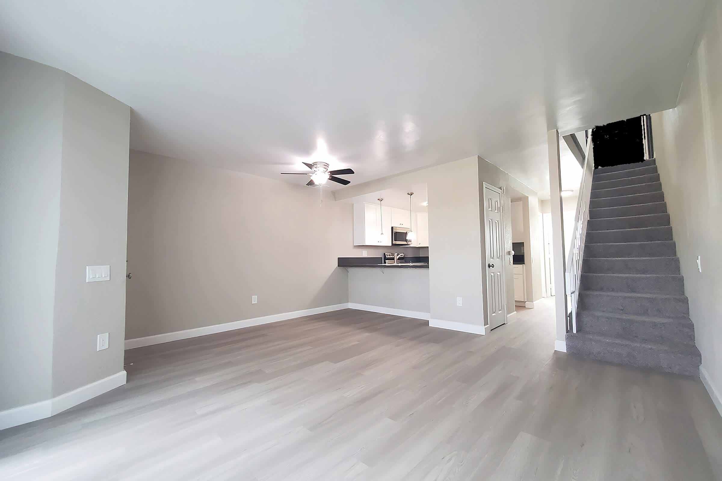 A spacious, modern room with light gray walls and a ceiling fan. The space features a kitchen area with white cabinetry visible in the background. To the right, there is a staircase leading to another level, and the floor is finished with a light wood-like material, creating a bright and airy atmosphere.