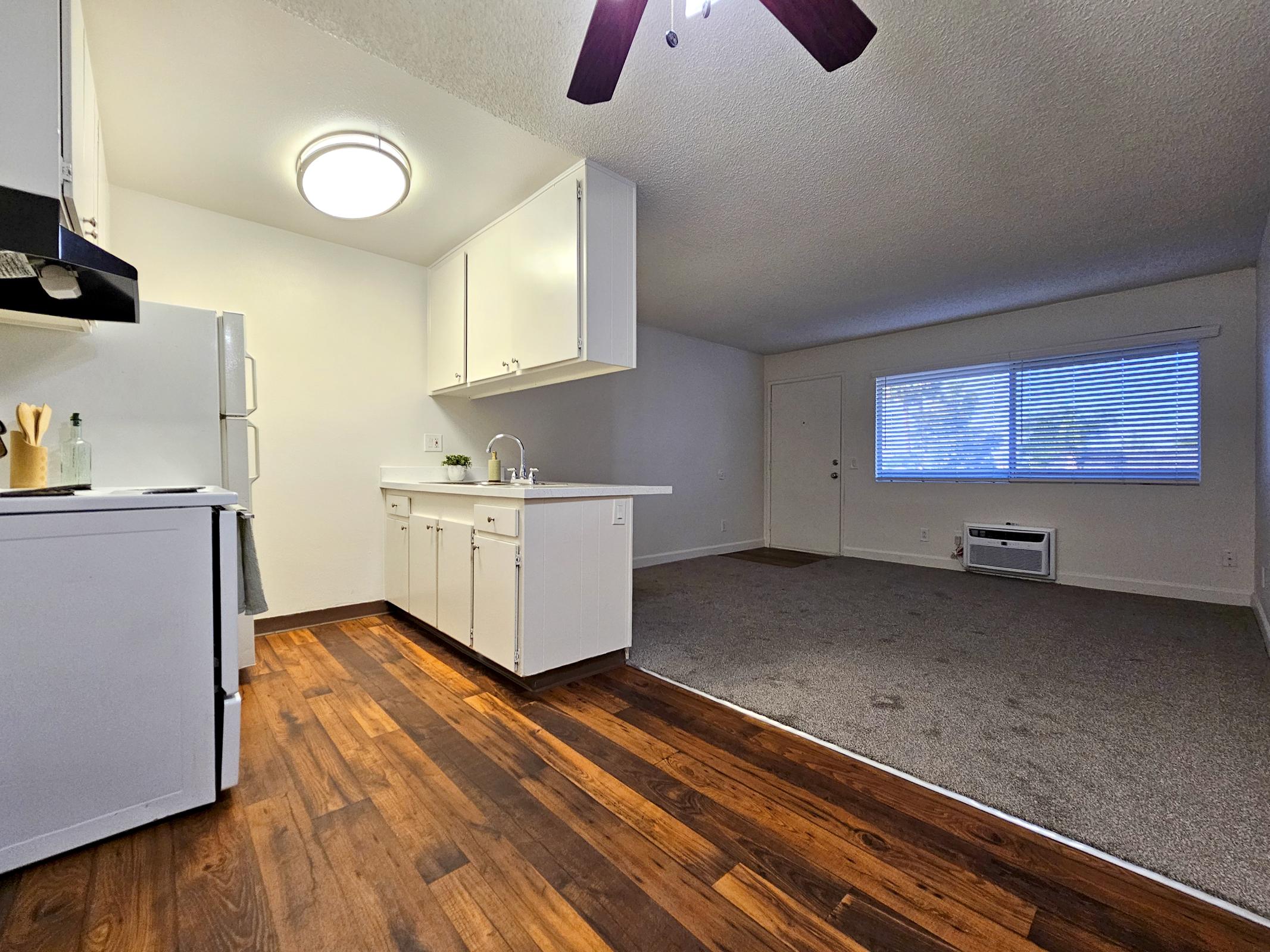A bright and open kitchen area with white cabinets, a sink, and a refrigerator visible. The hardwood floor contrasts with a carpeted living space adjacent to it, featuring a large window with blinds and an air conditioning unit on the wall. The room has a clean, modern look.