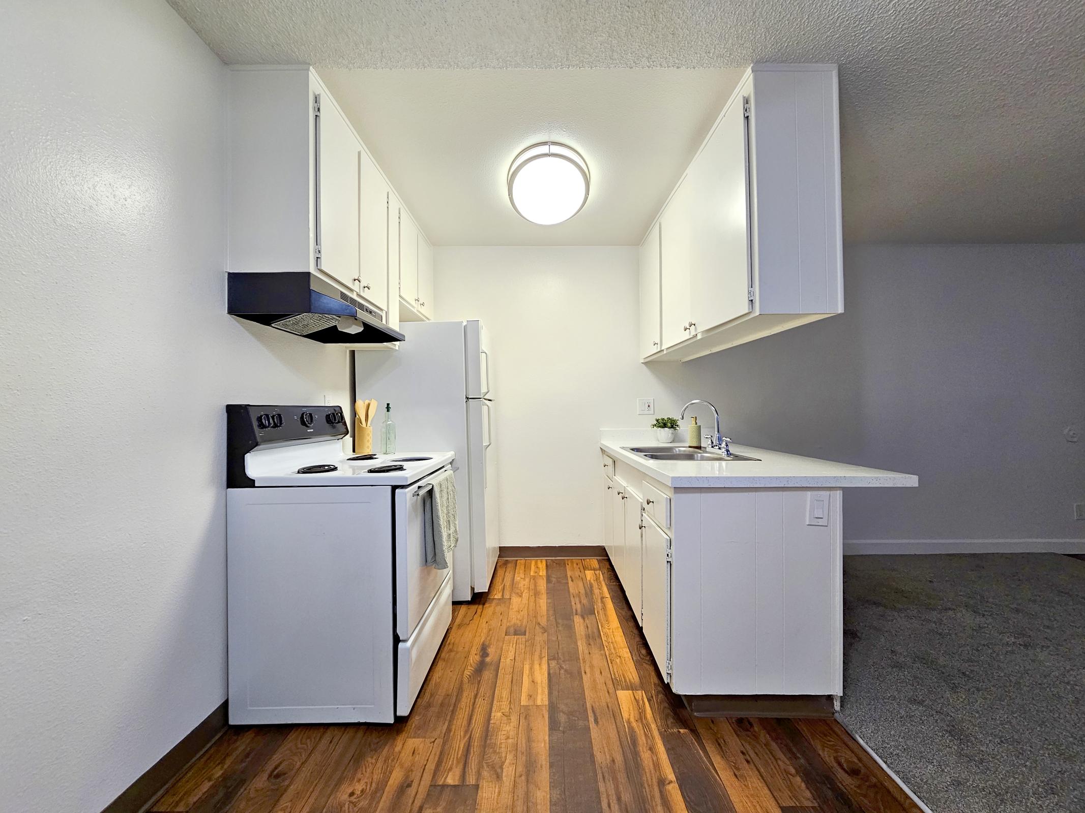 Modern kitchen with white cabinets, an electric stove, and a refrigerator. A countertop extends into an open layout, featuring a sink and small plant. The floor has warm wooden tones, contrasting with light walls and a bright ceiling light, creating a welcoming atmosphere.