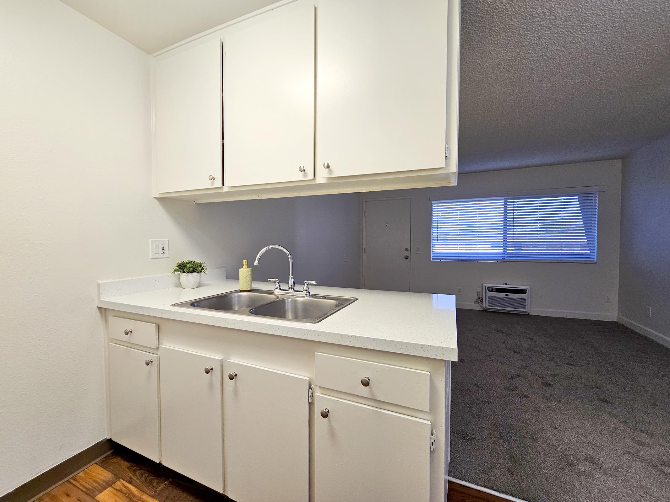 A clean, modern kitchen space featuring a white countertop with dual sinks, white cabinets, and minimal decor. In the background, a living area with a large window providing natural light, and light carpeting can be seen. The atmosphere appears bright and inviting.