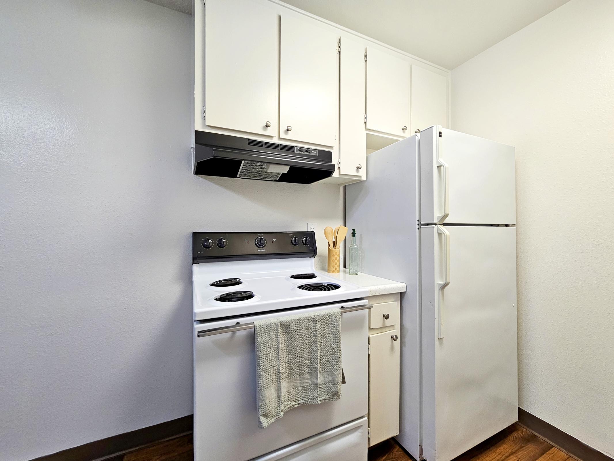 A bright kitchen featuring white cabinets, a white refrigerator, and a white stove with a hood. A kitchen towel hangs from the stove, and utensils are visible in a container. The walls are light-colored, and the flooring is dark wood, creating a clean and modern look.