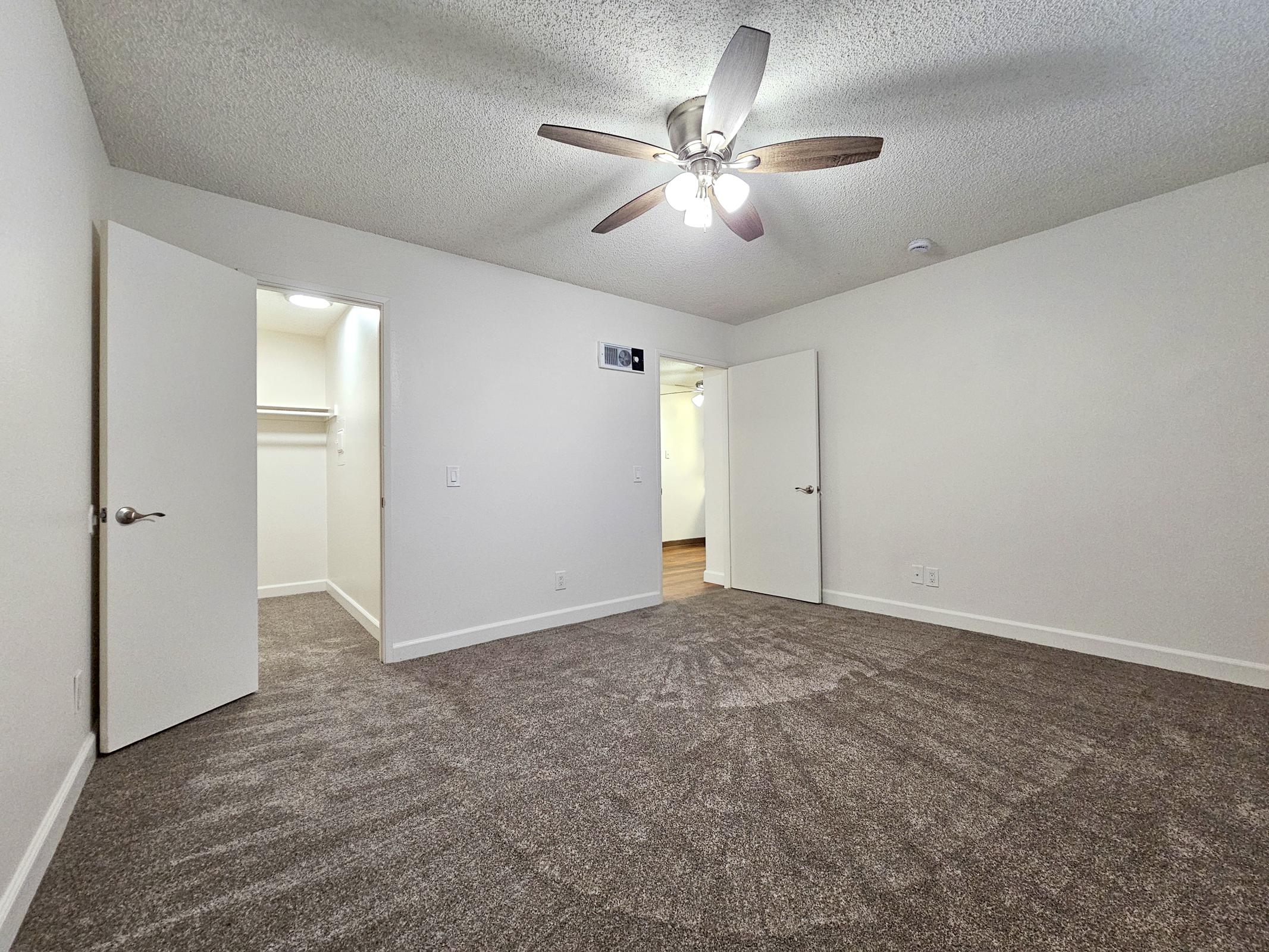A spacious room with light brown carpet, a ceiling fan, and a neutral color scheme. Two white doors lead to a closet and another area, while the room's layout offers ample space for furniture. The lighting is bright, enhancing the airy feel of the space.