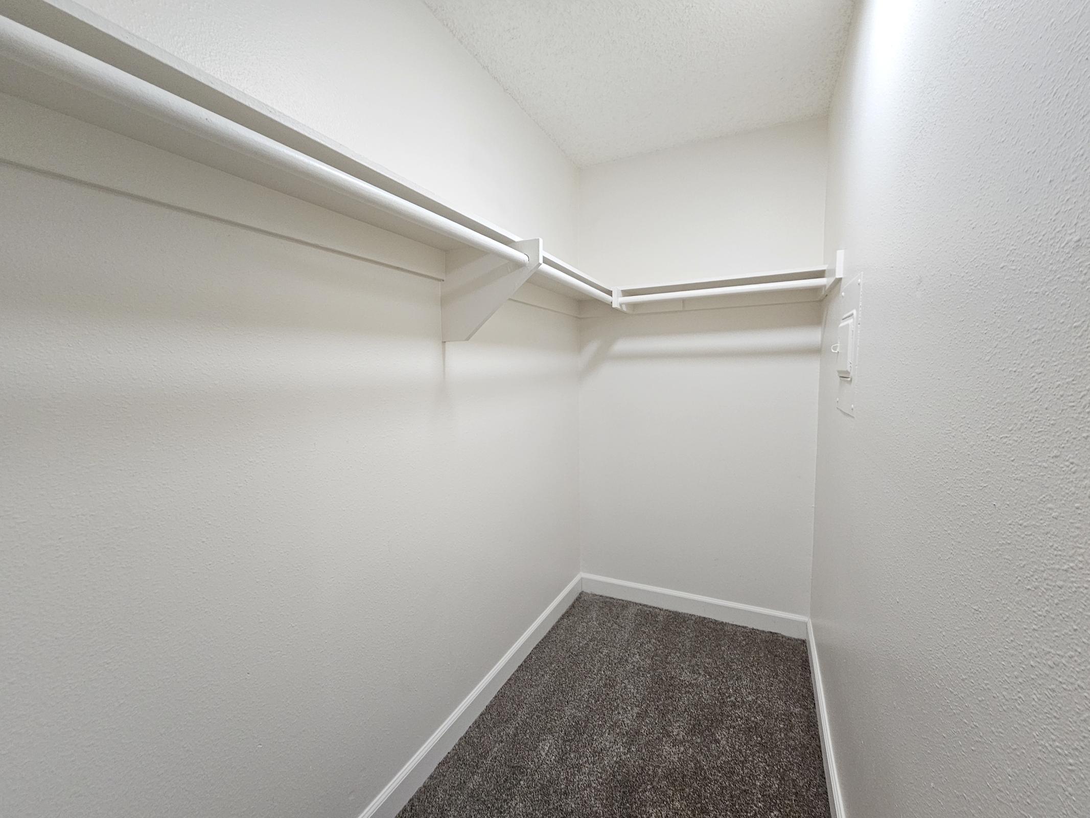 Empty closet with white walls and shelving. The floor is covered in carpet, and there are two shelves on one side. The lighting is bright and there are no visible items or decorations in the space.