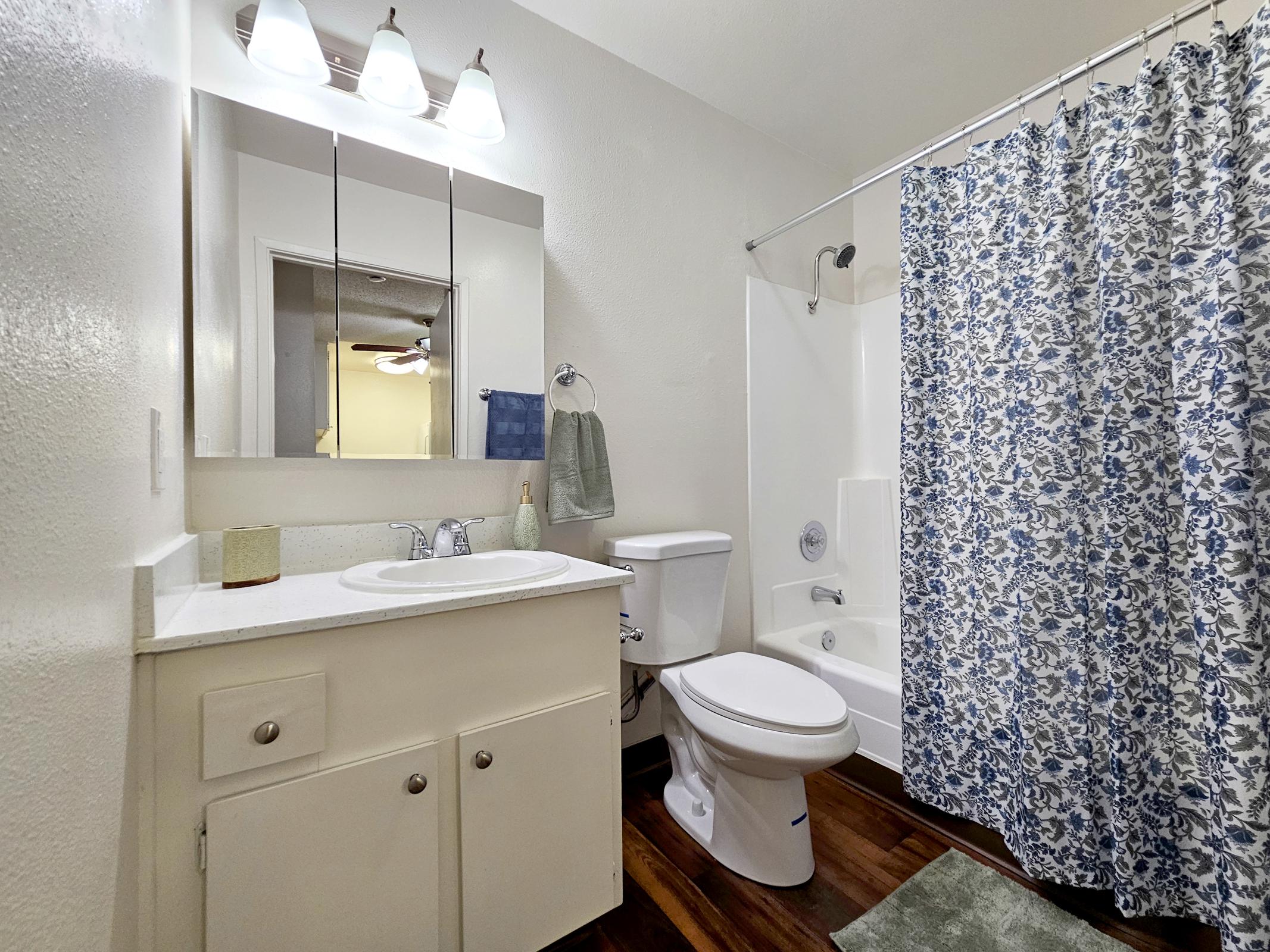 Bathroom interior featuring a white sink and cabinet, a toilet, and a bathtub with a shower curtain displaying a blue floral pattern. A mirror and wall-mounted light fixtures are above the sink, and there is a small towel hanging on a rack. The flooring is wooden, adding warmth to the space.