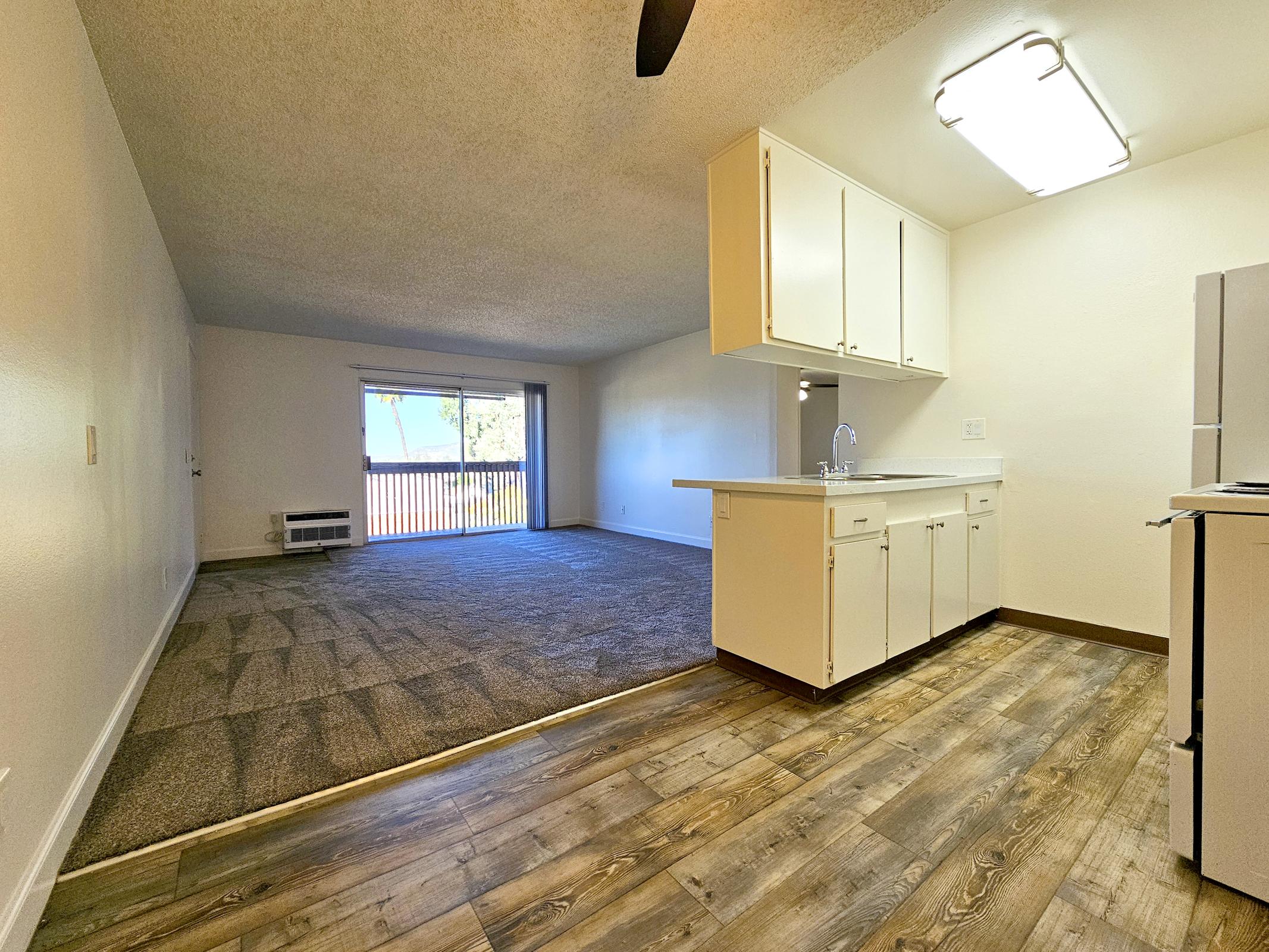 Looking into a spacious, well-lit room featuring a kitchen with white cabinets and a countertop. The living area has beige carpet and a large window with a balcony view. Natural light fills the space from a ceiling light fixture, creating an inviting atmosphere.