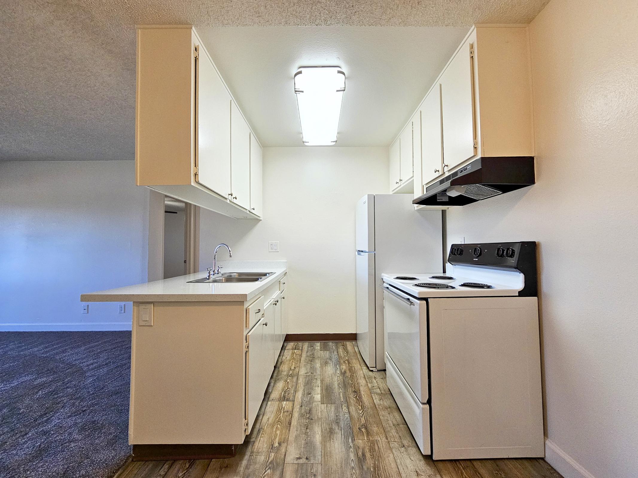 A modern kitchen featuring white cabinetry, a stainless steel sink, a refrigerator, and a black stove with an oven. The countertops are light-colored, and the flooring is a warm, wood-like material. The space is well-lit with overhead lights and has a carpeted area visible in the background.