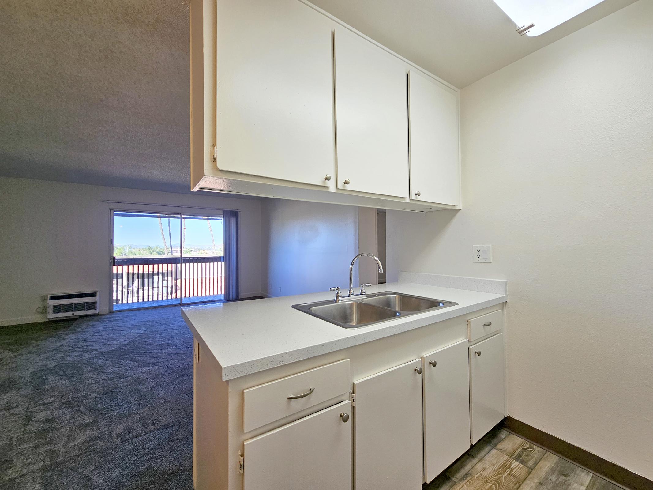 A small kitchen area featuring white cabinets, a double sink, and a countertop. In the background, a view of a spacious, carpeted living area with a large window looking out. Natural light fills the space, creating an inviting atmosphere.