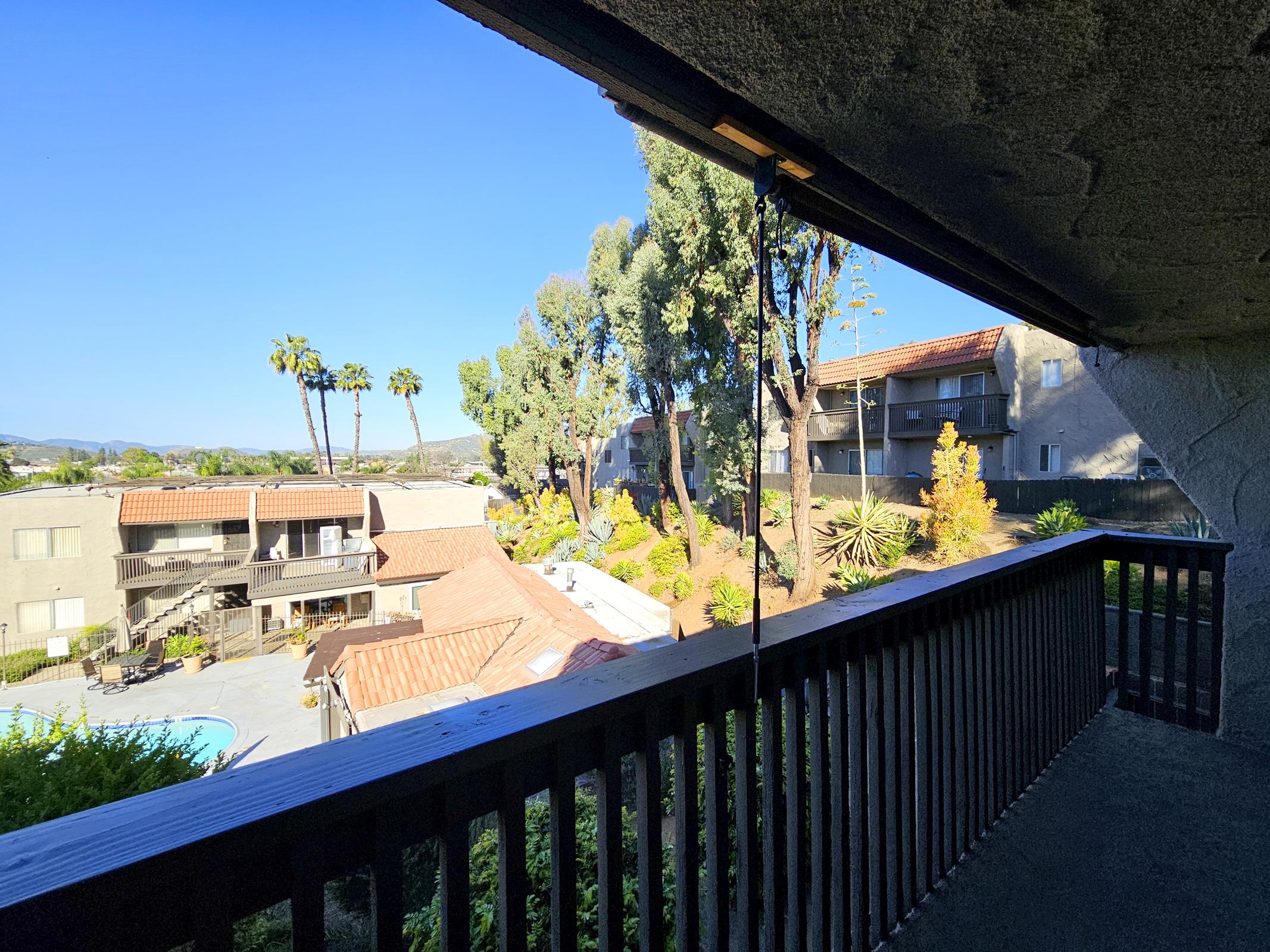 View from a balcony overlooking a landscaped area with palm trees and other greenery. In the foreground, there is a wooden railing, while the background features several apartment buildings and a clear blue sky. The scene is bright and showcases a sunny day.