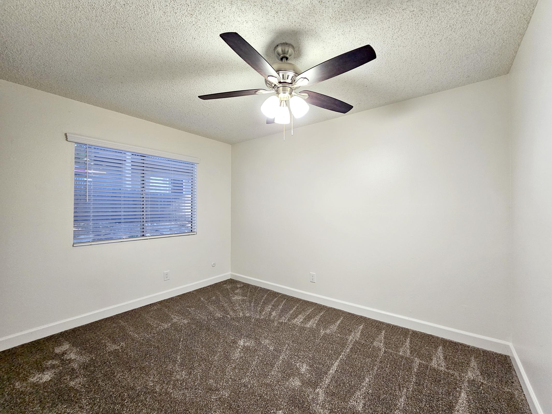 A spacious room with light-colored walls and a ceiling fan. The floor is covered in plush, brown carpet, and there is a window with blinds allowing natural light to enter. The room is empty, creating a minimalist and clean appearance.
