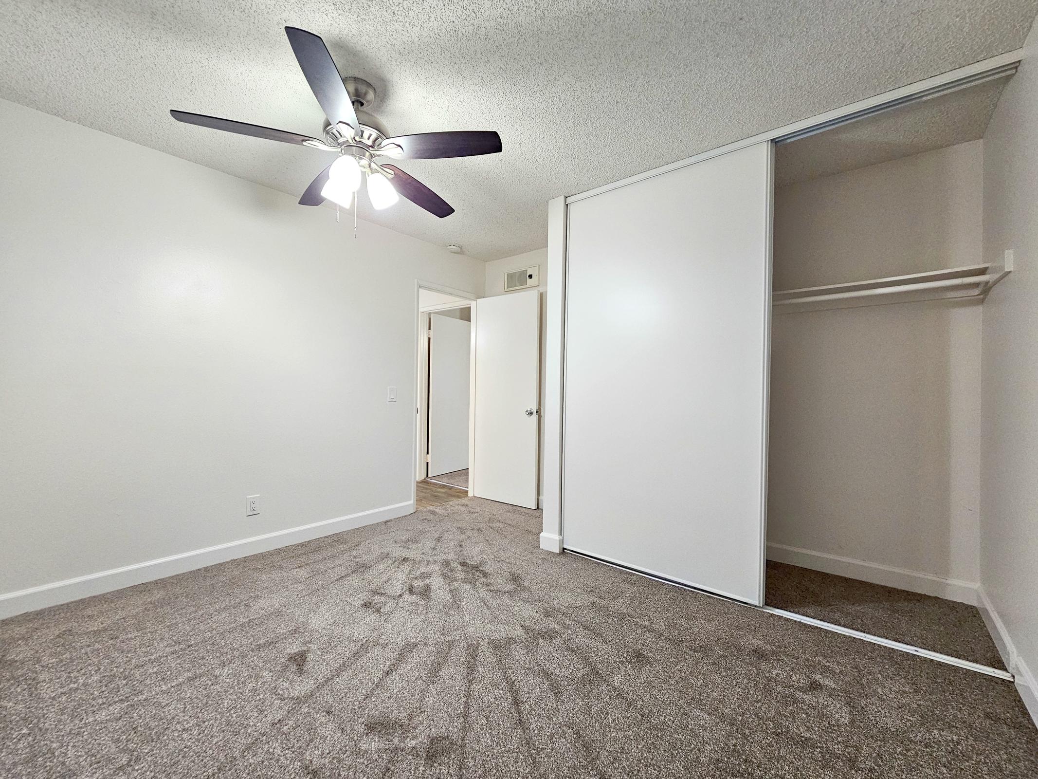 A spacious, well-lit room featuring light carpet, a ceiling fan with five blades, and a mirrored sliding closet door. The walls are painted in a neutral tone. A door leads to another room, adding to the open feel. The room is ideal for various uses, such as a bedroom or office.
