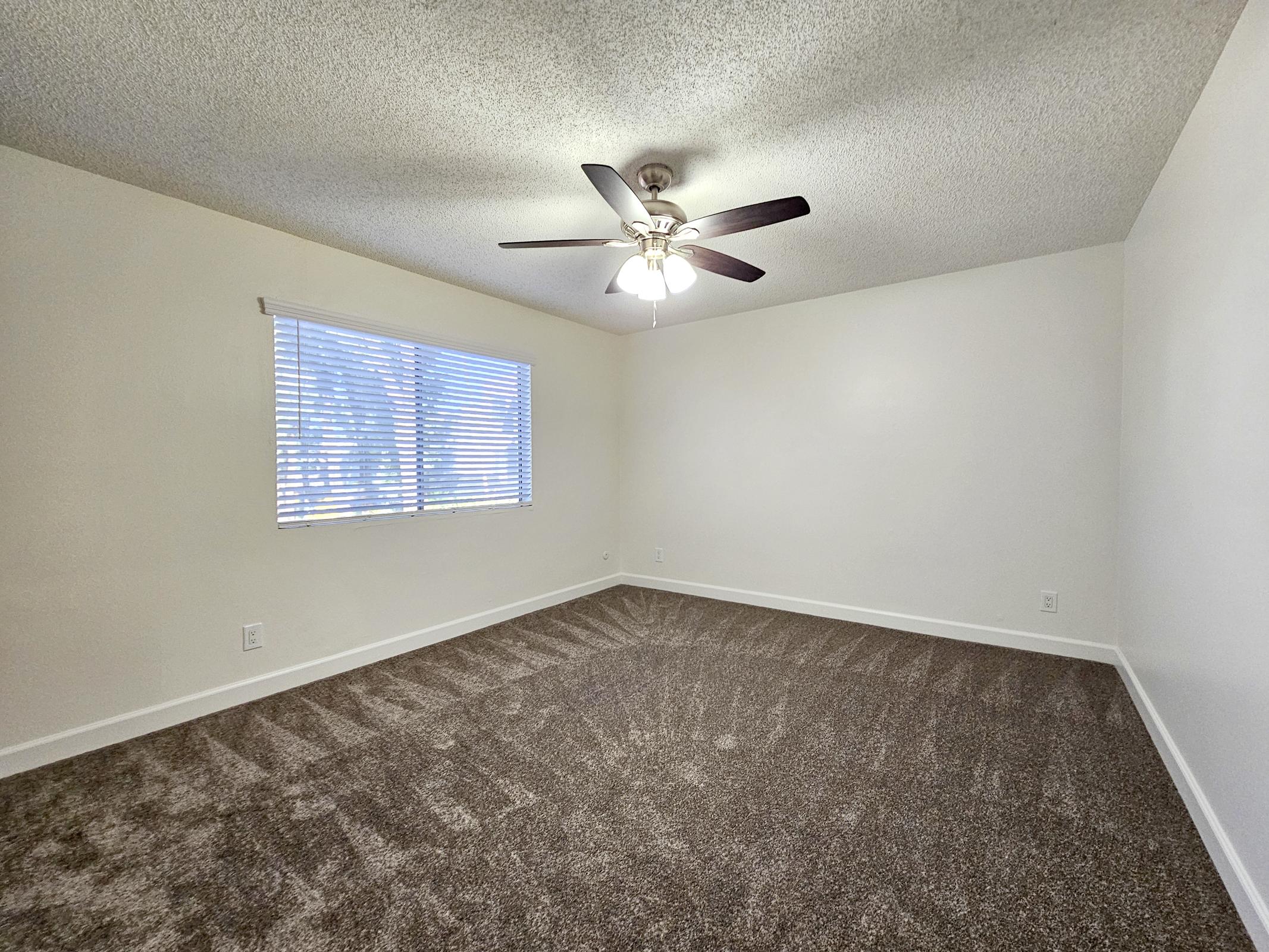 A spacious, empty bedroom featuring light-colored walls and a textured ceiling. The room has a ceiling fan with three lights, and a window with blinds that lets in natural light. The floor is covered with soft, brown carpet, creating a warm and inviting atmosphere.