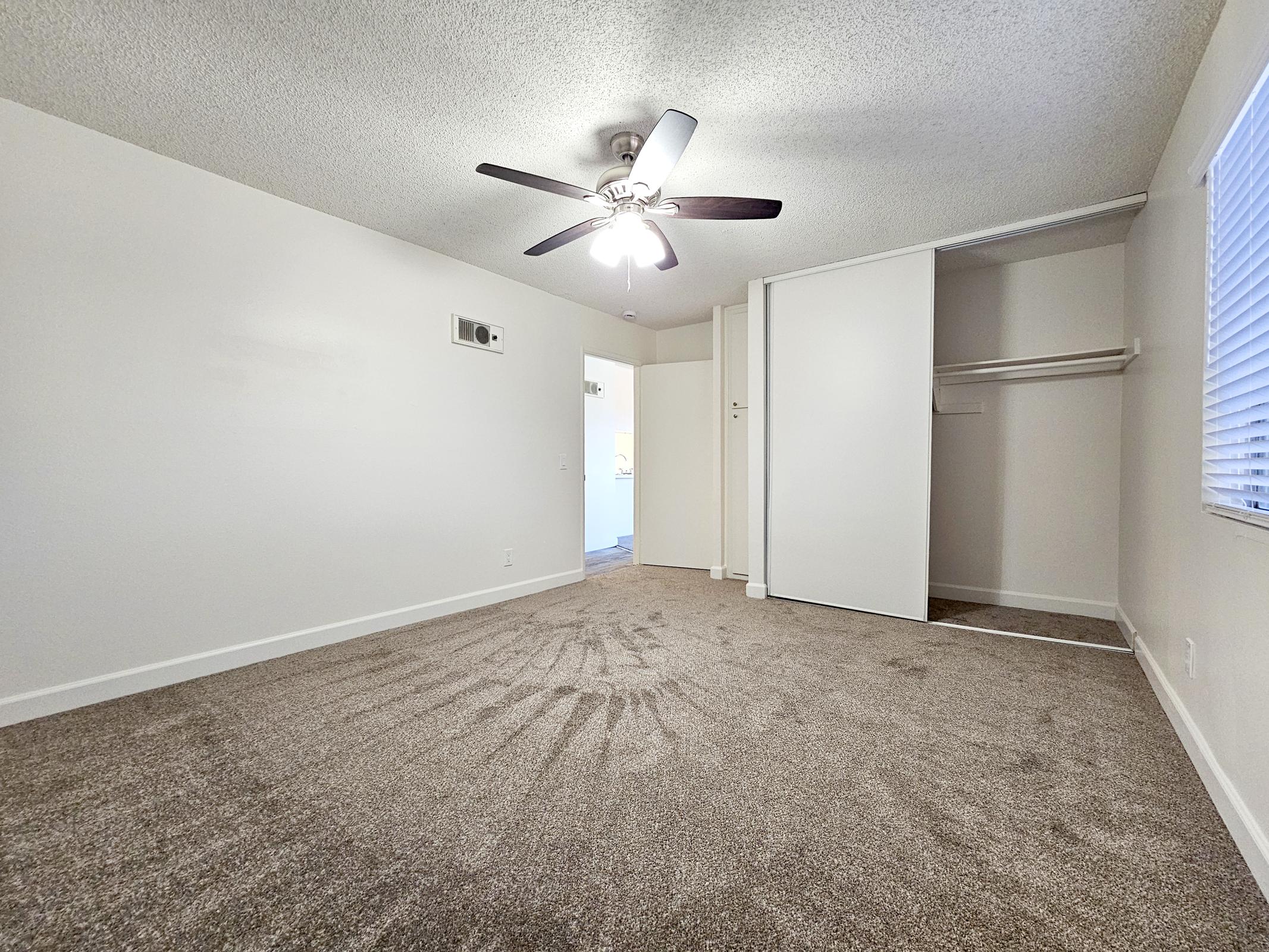 A well-lit, empty room with a ceiling fan, light gray walls, and a window. The floor is covered in carpet with a circular pattern. There is a closet with sliding doors on one side, and a small air vent on the wall. The space appears clean and ready for moving in.