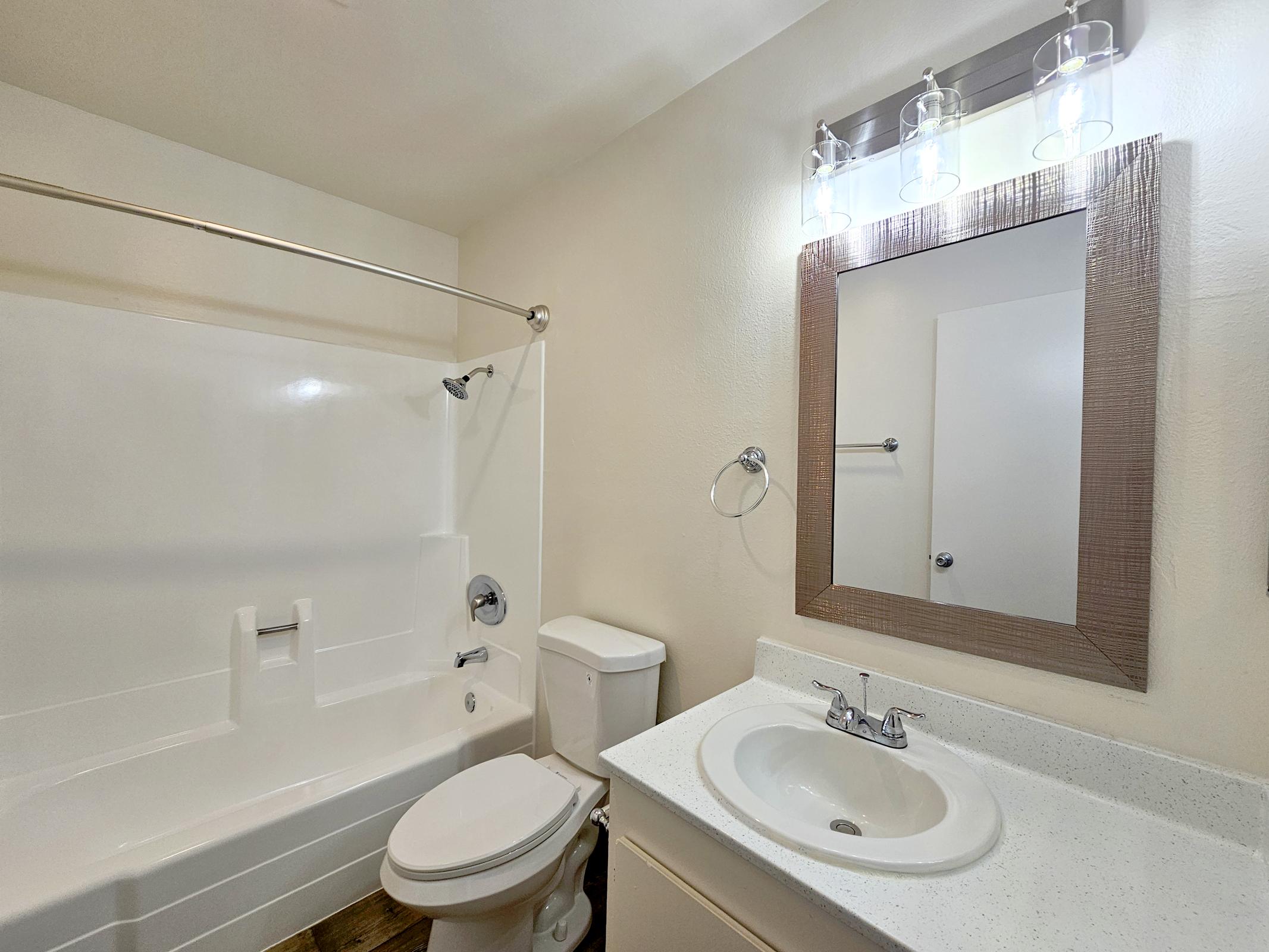 A clean, well-lit bathroom featuring a white tub with a shower curtain, a modern toilet, and a sink with a faucet. A rectangular mirror is above the sink, surrounded by a stylish frame, while three light fixtures provide illumination. The walls are painted a light color, enhancing the fresh ambiance.