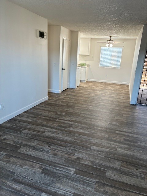 A spacious and bright interior of an apartment featuring light-colored walls, new laminate flooring, and a ceiling fan. On the right, there is a doorway leading to a kitchen area with cabinets and a window, while the left side shows a hallway leading to additional rooms.