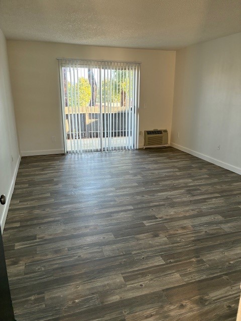 Empty living room featuring a large window with vertical blinds, allowing natural light to enter. The floor is covered with gray laminate flooring, and an air conditioning unit is visible on the right. The walls are painted a light color, creating a spacious and bright atmosphere.