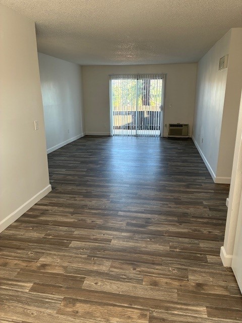 A spacious and airy living room with light-colored walls and wooden flooring. Large sliding glass doors lead to a balcony, allowing natural light to stream in. The room is empty, creating an open and inviting atmosphere, with an air conditioning unit visible in the corner.