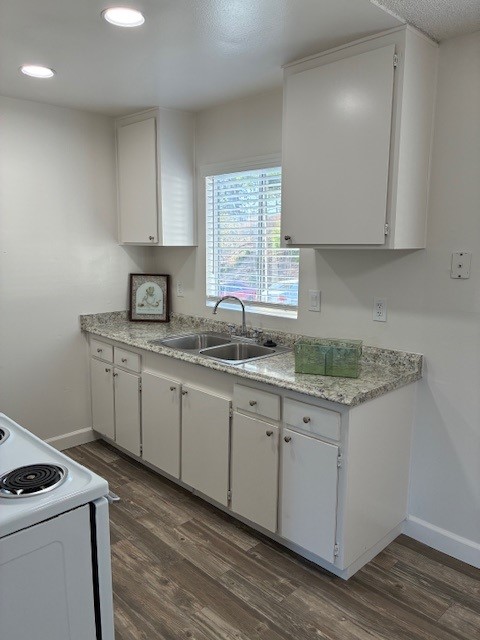 A modern kitchen featuring white cabinets and a granite countertop. A double sink is positioned under a window with natural light. There is a framed picture on the counter and a glass container. The floor is made of wooden planks, and a stovetop is visible in the foreground.