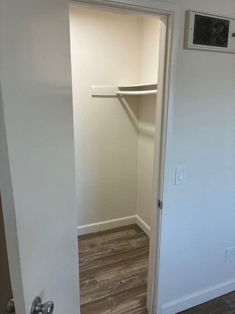 A small, empty closet with a white door partially open. Inside, there's a shelf along the back wall and light-colored walls. The floor is covered with light wood-like laminate, and there is an air vent on the wall above the door. The overall space is well-lit and clean.