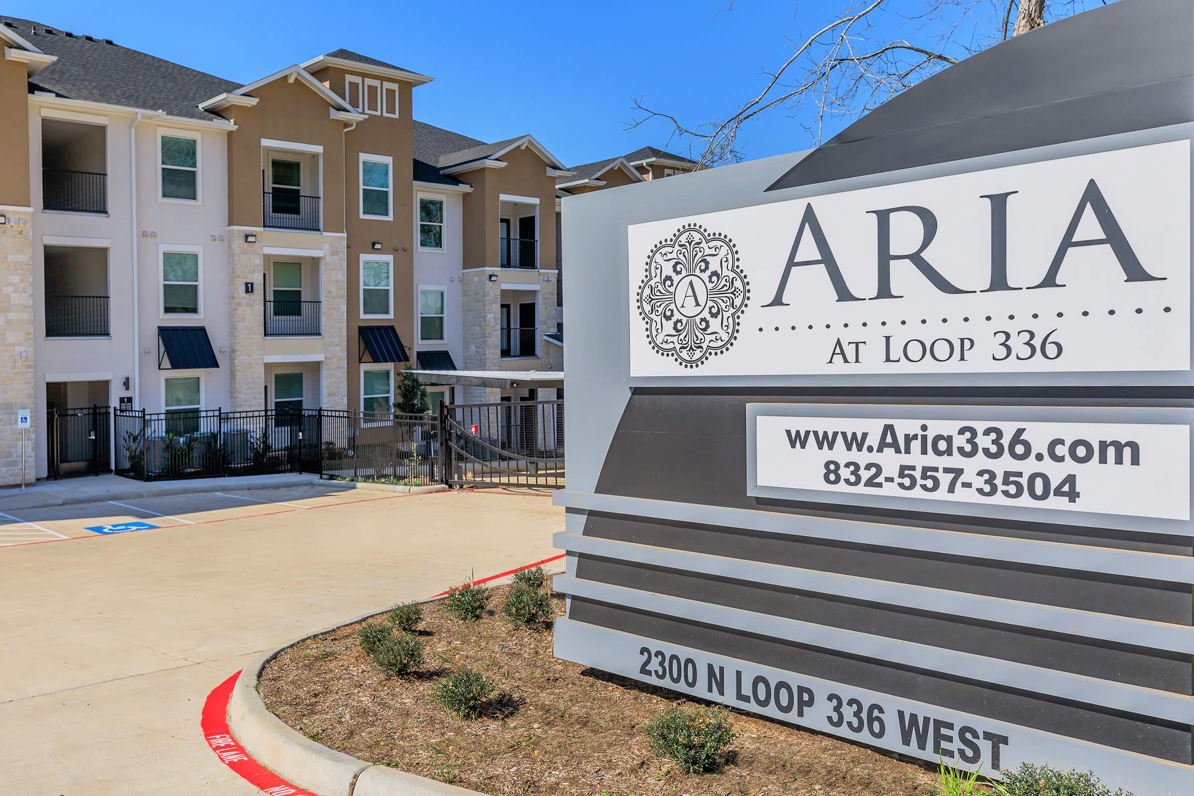 Sign for Aria at Loop 336, featuring the name and contact information. In the background, a modern apartment building with balconies and contemporary architecture is visible. The area is well-maintained with landscaping, and the sign includes directional information and a parking space indicator.