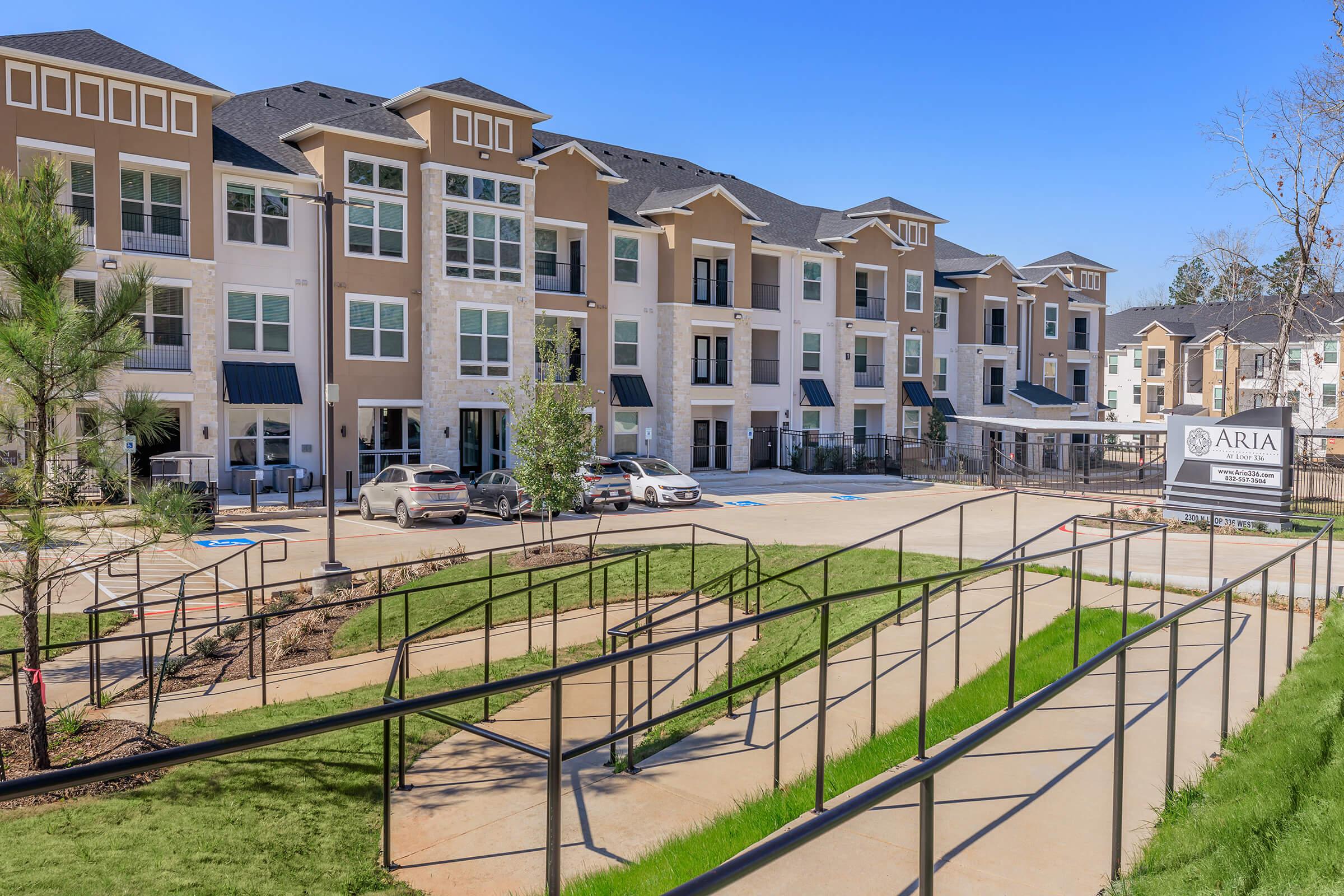 A modern apartment complex featuring multiple buildings with a well-maintained landscape. The foreground includes a sloped pathway with railings leading up to the entrance. Several parked cars are visible in the lot, alongside a sign displaying the name "ARIA." The sky is clear and blue, indicating a sunny day.