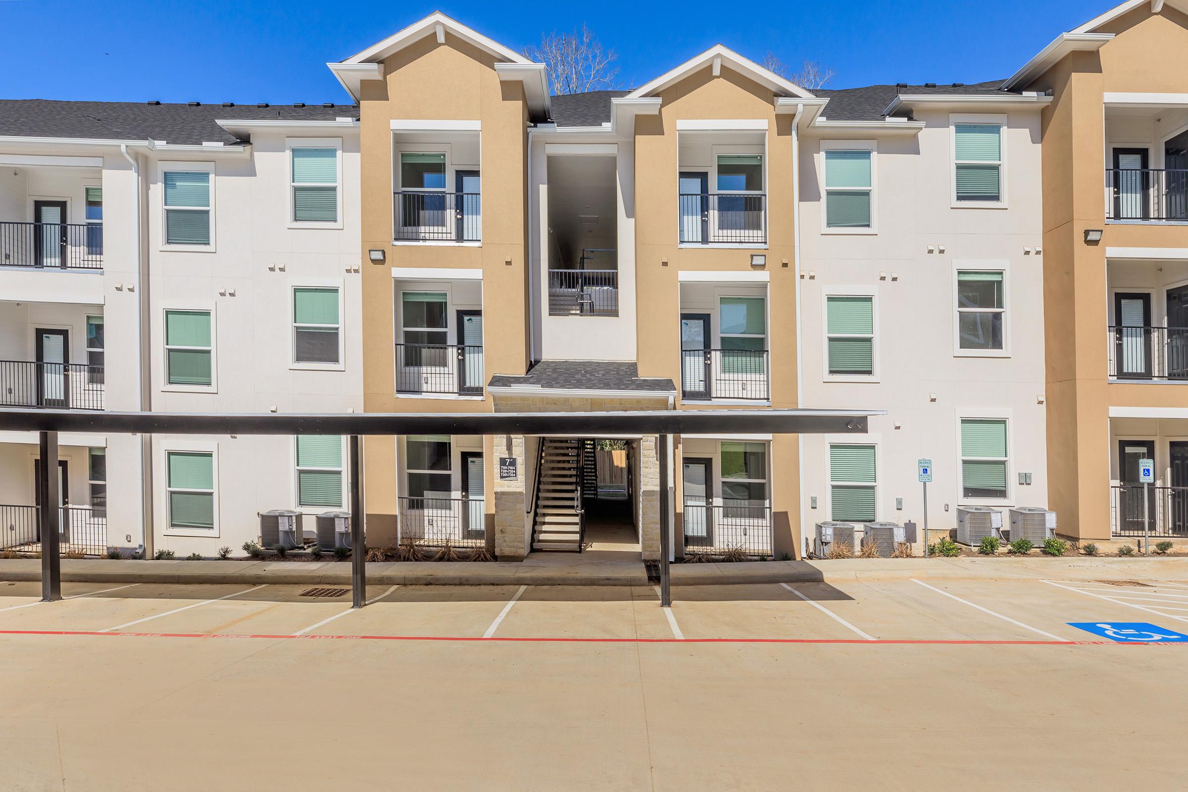 View of a modern apartment building with two stories, featuring a light exterior and multiple windows. The entrance is centrally located with stair access. In the foreground, there is a parking area with designated spots and a covered parking structure. The sky is clear and blue, providing bright lighting.