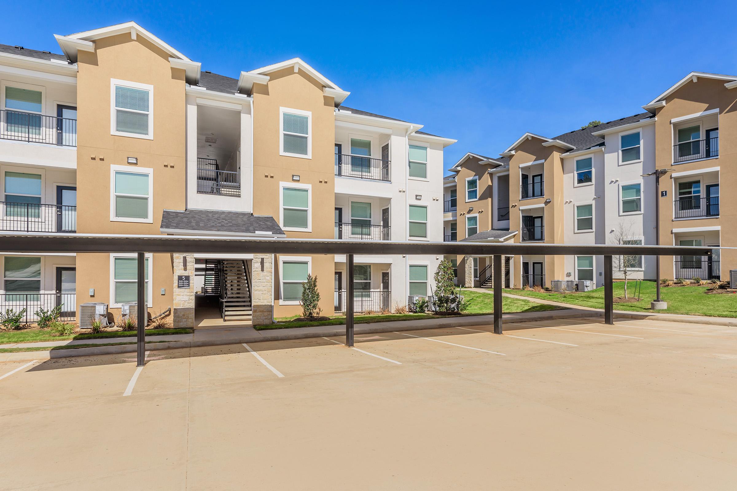 Modern multi-unit apartment buildings with beige exteriors and balconies. A parking area with covered spaces in the foreground and green landscaping surrounding the buildings. The sky is clear and blue, indicating a bright day.