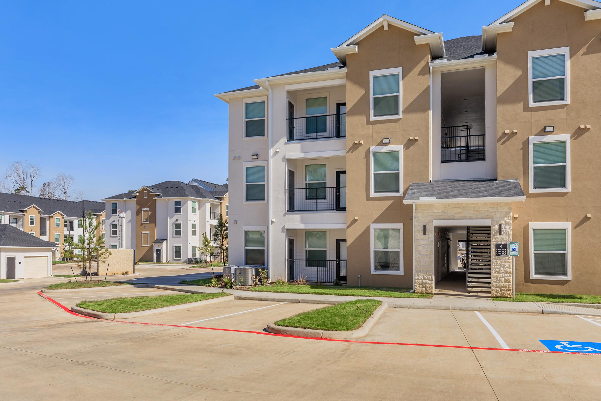 A modern apartment complex featuring multiple buildings with light-colored façades. The foreground shows a paved parking area with designated parking spaces, and a well-maintained lawn. Clear blue sky above enhances the bright appearance of the development.