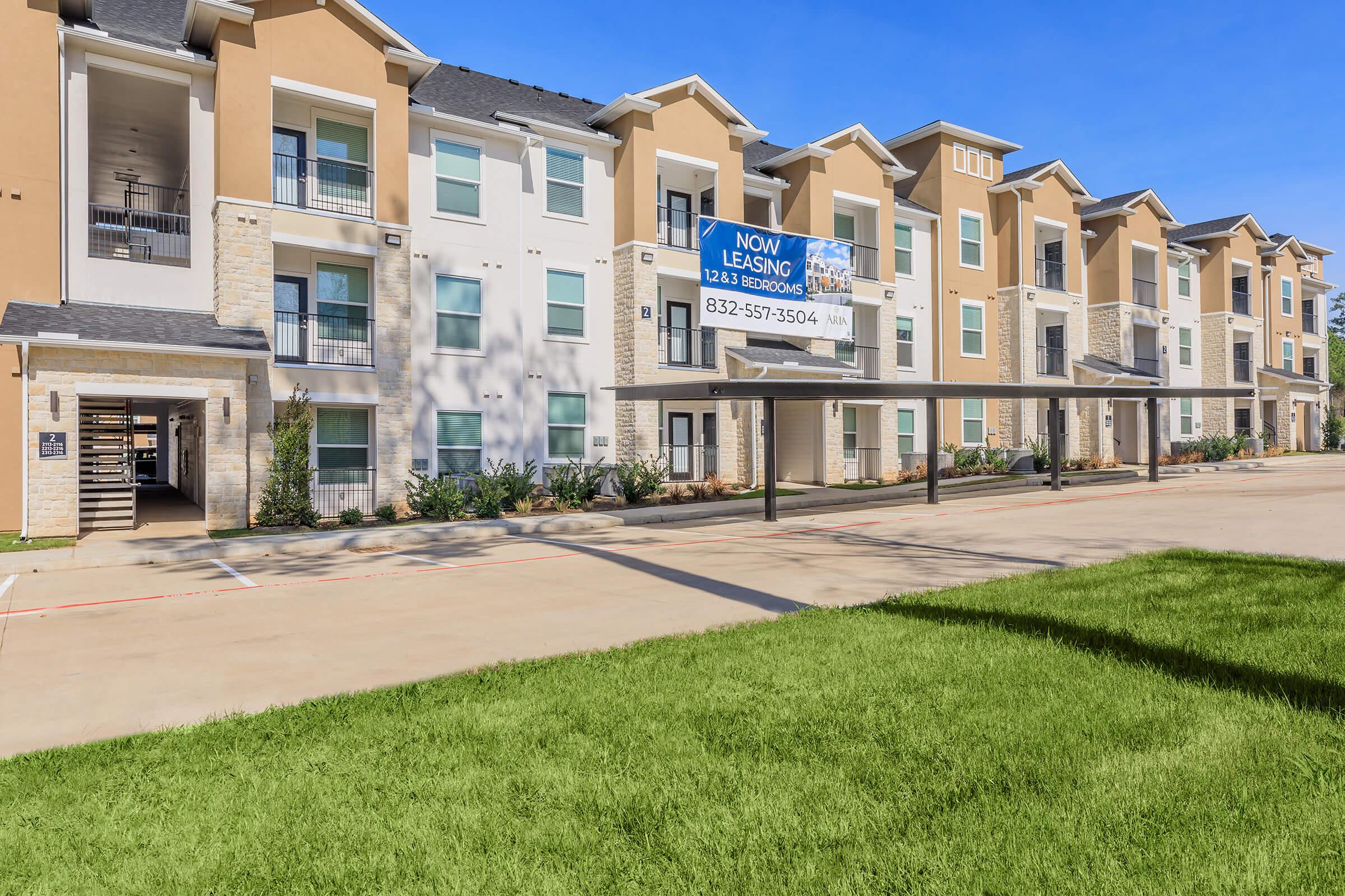 A modern apartment building with multiple levels, featuring a large "Now Leasing" banner. The exterior is a mix of light colors with stone accents. The parking area is covered, and there is a well-maintained green lawn in front.