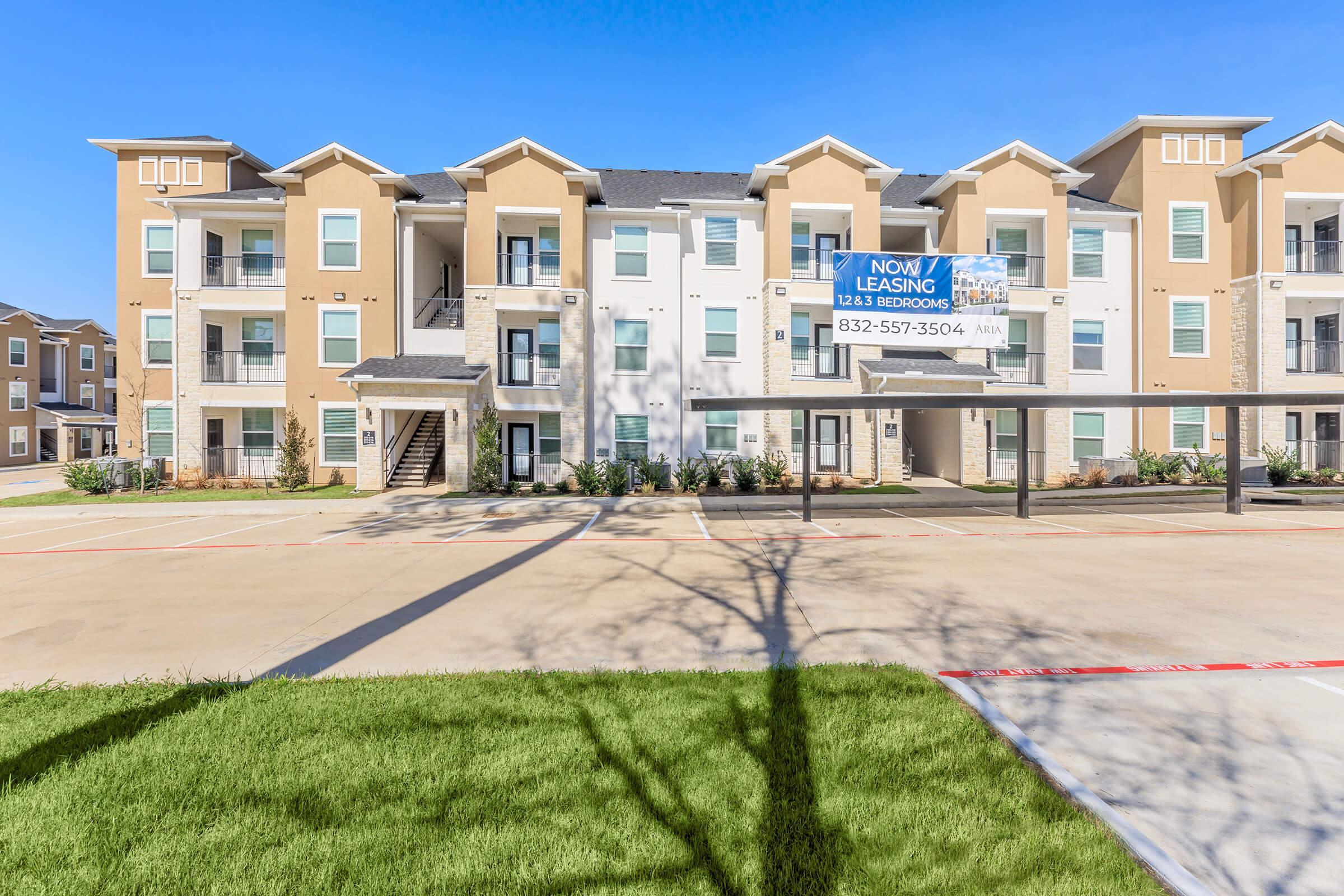 An apartment complex with modern architecture, featuring a two-tone color scheme of beige and white. A large leasing sign is prominently displayed in front of the building, which has multiple levels and balconies. The surrounding area includes a freshly paved parking lot and well-maintained grass.