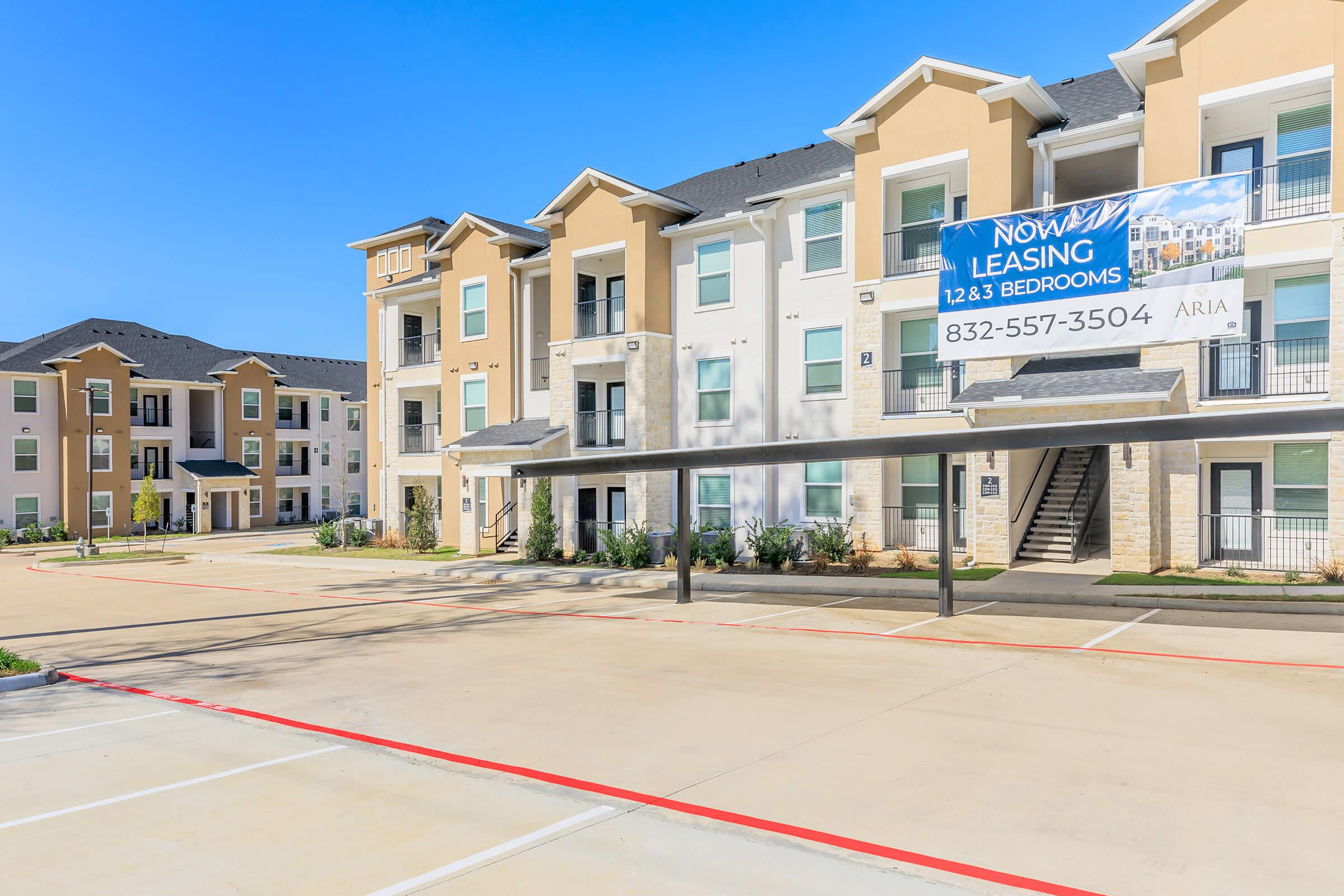 A modern apartment complex featuring two buildings with multiple units. The foreground shows a parking area with marked spaces. A prominent "Now Leasing" sign highlights available 1, 2, and 3-bedroom apartments. The sky is clear and blue, enhancing the fresh look of the property.