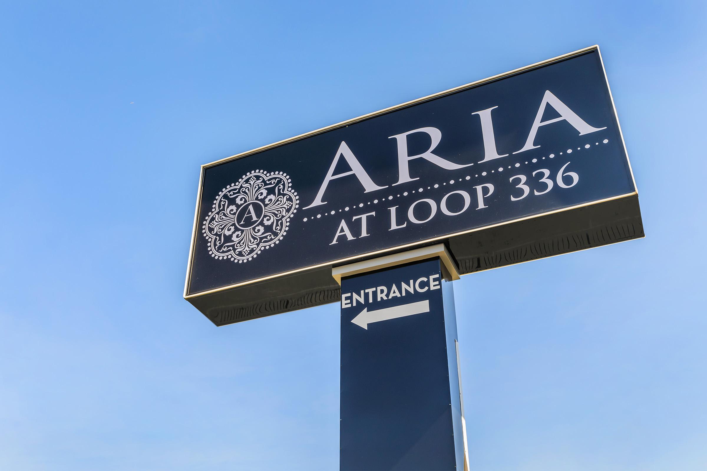 Sign for Aria at Loop 336 with a decorative logo, displaying the word "ARIA" in large letters and directing towards an entrance with an arrow pointing to the left. The sign is set against a clear blue sky.
