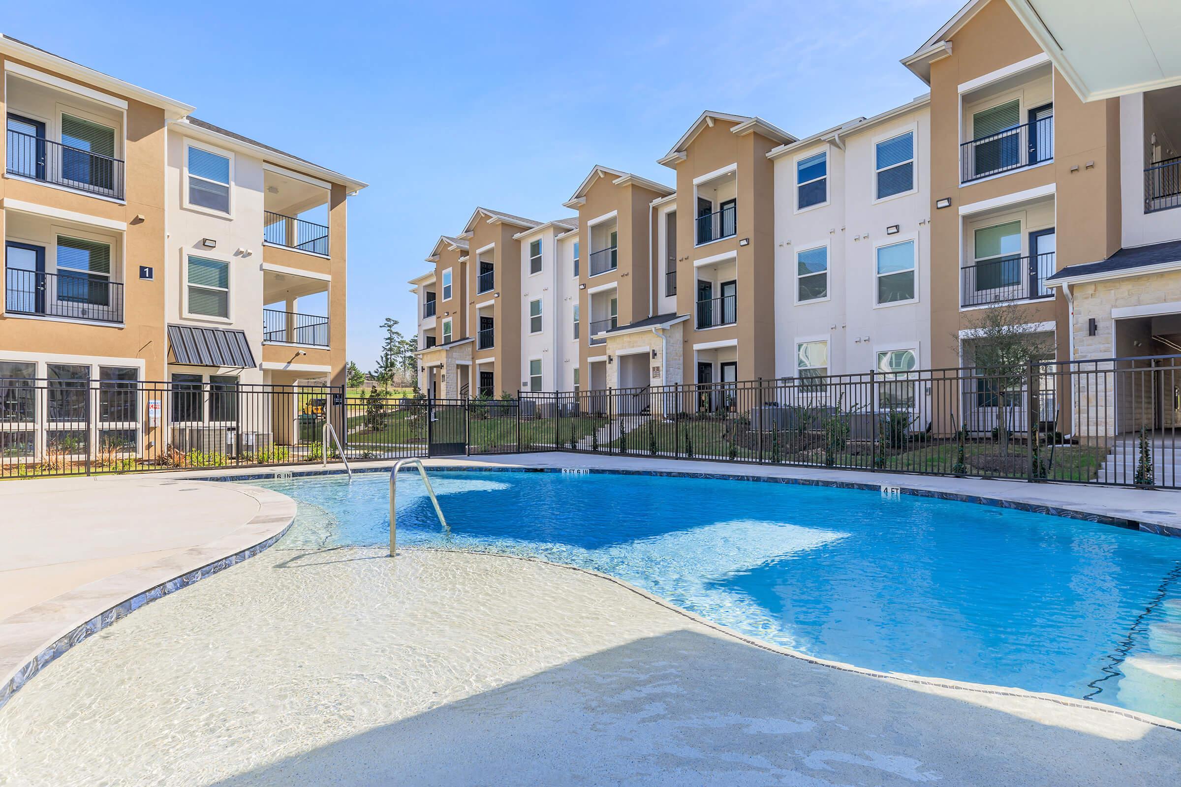 Swimming pool surrounded by modern apartment buildings, featuring a smooth surface and a shallow entry area. The pool is enclosed by a black fence, with landscaped greenery in the background and clear blue skies above.