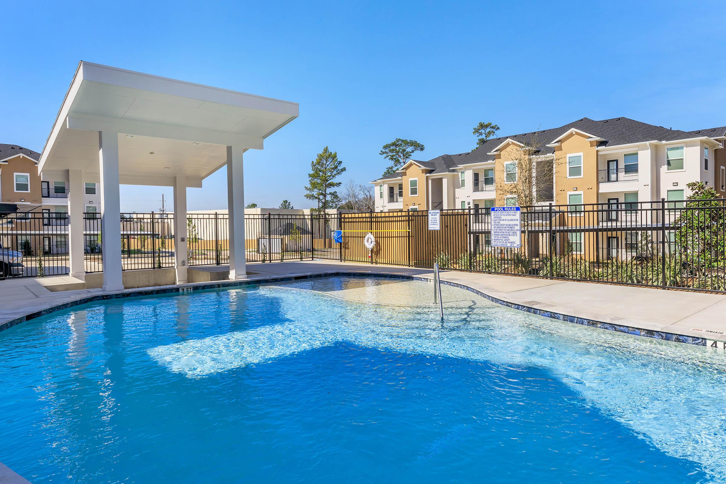 A clear blue swimming pool surrounded by a well-maintained deck. In the background, there are modern apartment buildings with balconies. A shaded gazebo is nearby, and the area is well-fenced with a few trees lining the space, creating a relaxing outdoor environment.