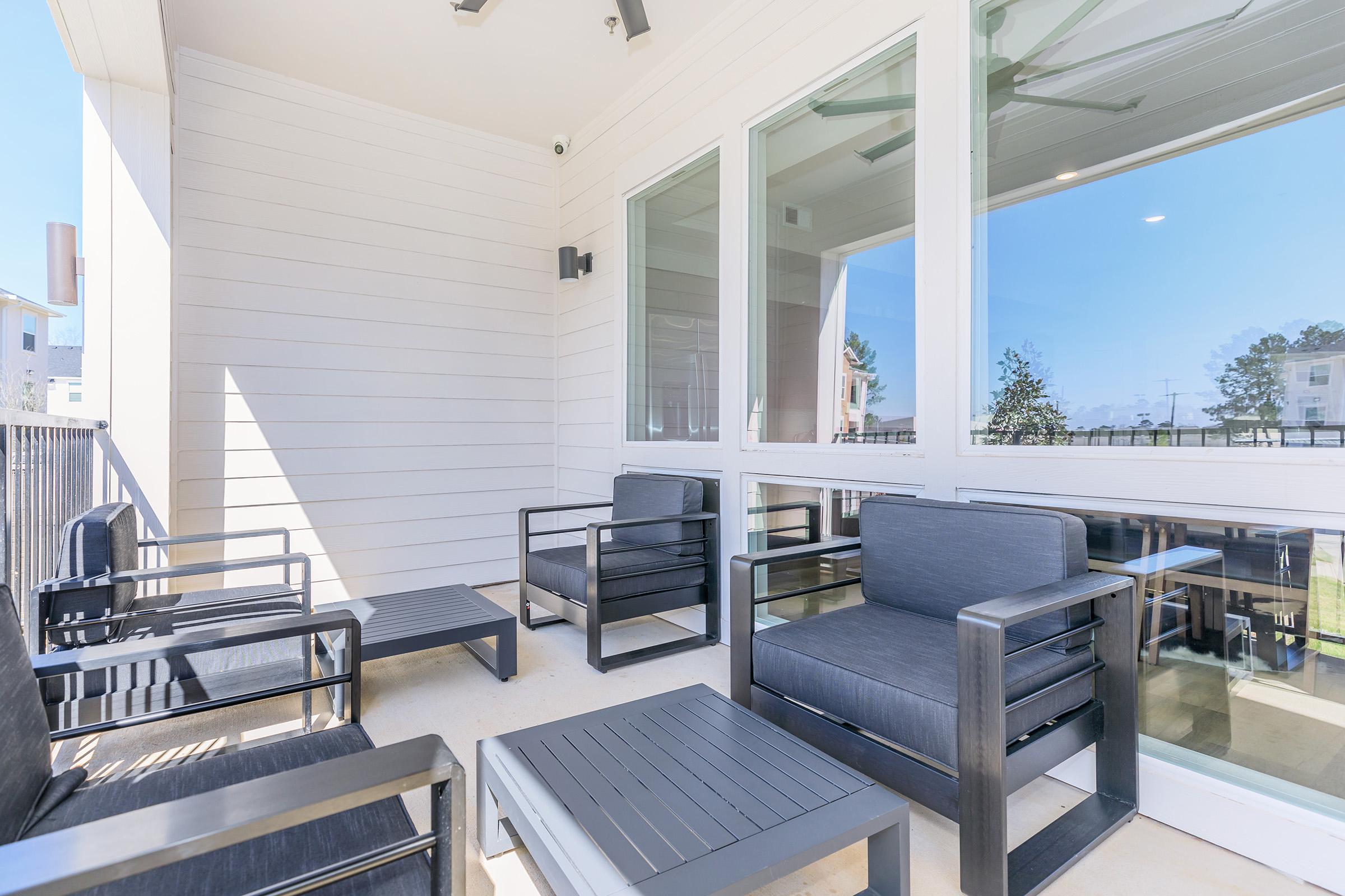 A modern outdoor balcony with several black seating chairs and a small coffee table. Large windows provide a view of the outdoors, featuring a clear blue sky and greenery. The space is well-lit and designed for relaxation or socializing.