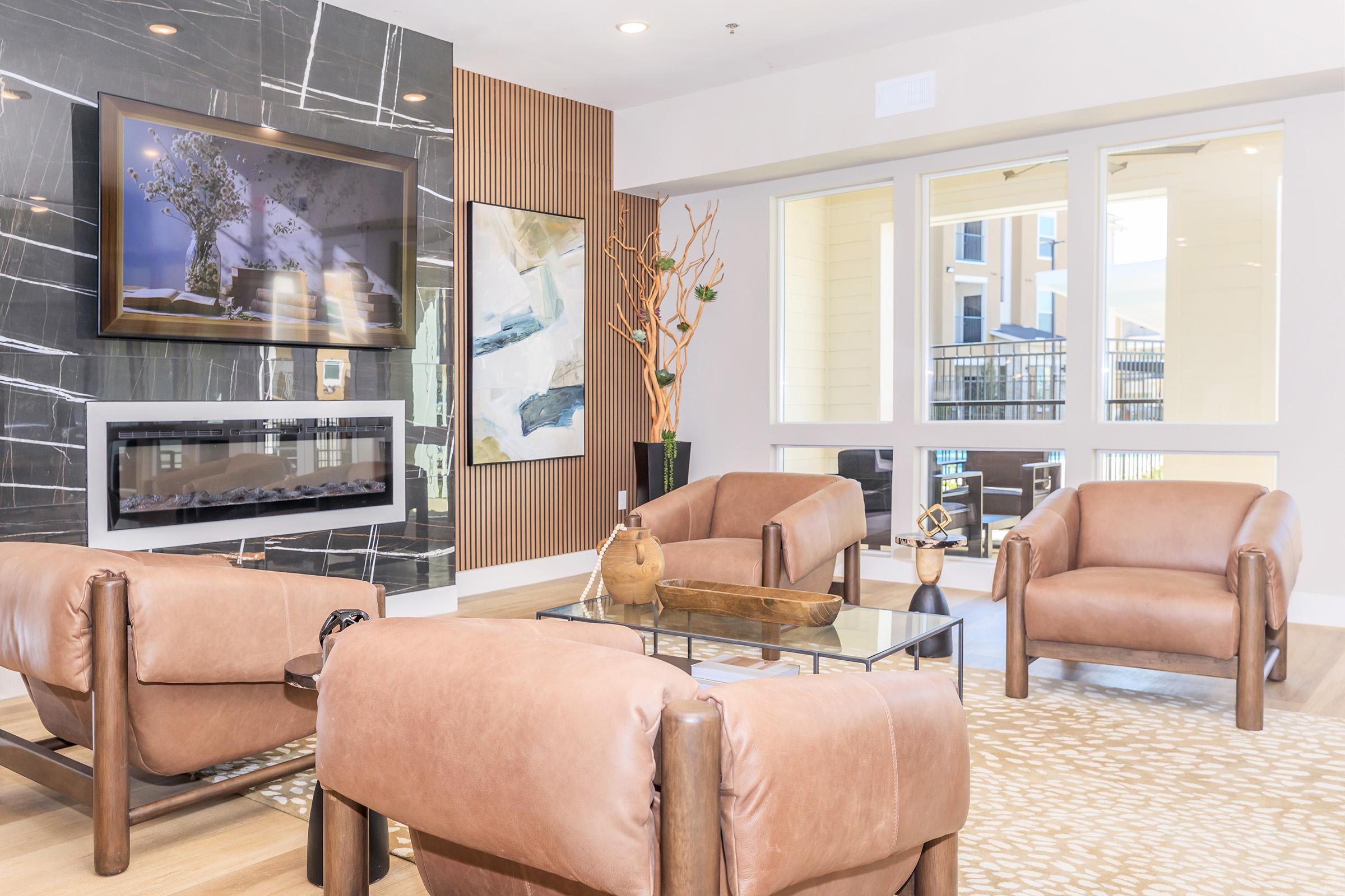 A modern living area featuring four tan leather chairs arranged around a glass coffee table. The wall behind has a sleek black and white marble design with framed artwork. There is a decorative plant and a fireplace below a large window, allowing natural light to brighten the space.