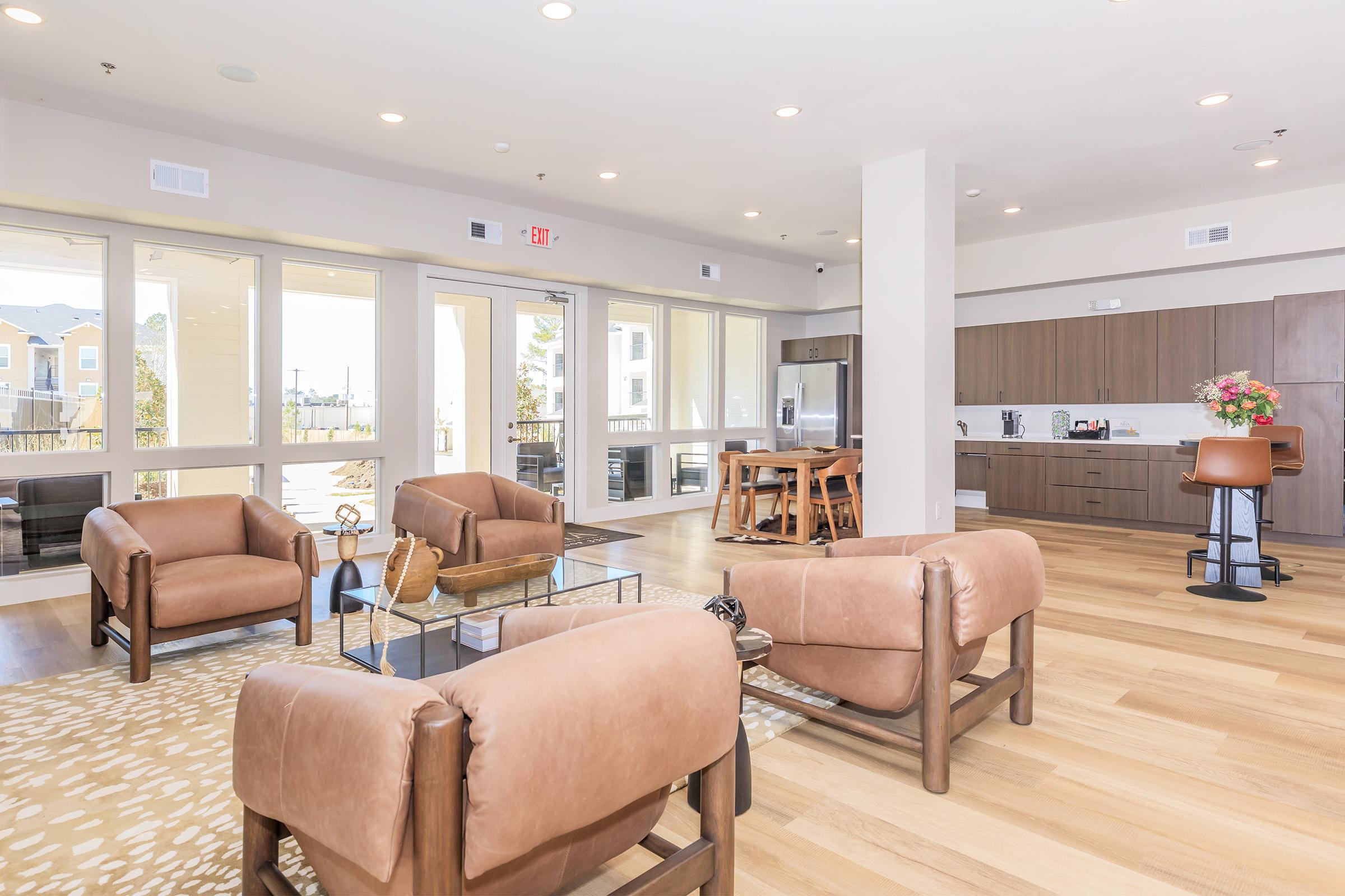 A modern living room with large windows allowing natural light. It features two brown leather armchairs, a coffee table, and a dining area with a wooden table and chairs. The kitchen is visible in the background, with contemporary cabinetry and appliances. The floor has light-colored wood and a decorative rug.