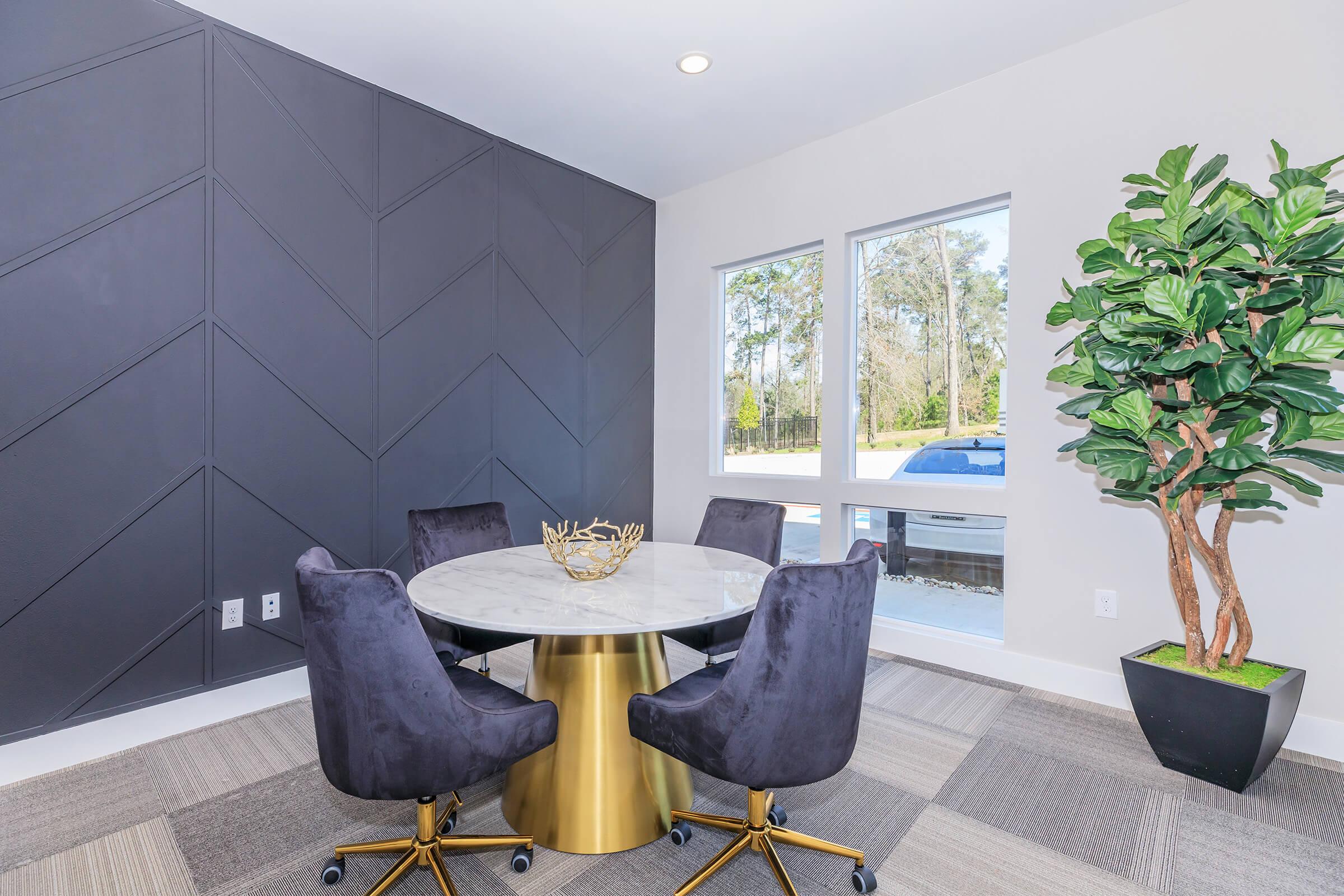 A modern dining area featuring a round marble table surrounded by four black upholstered chairs. The room has a stylish gray chevron accent wall and a large window letting in natural light. A decorative plant in a black pot adds a touch of greenery to the space.