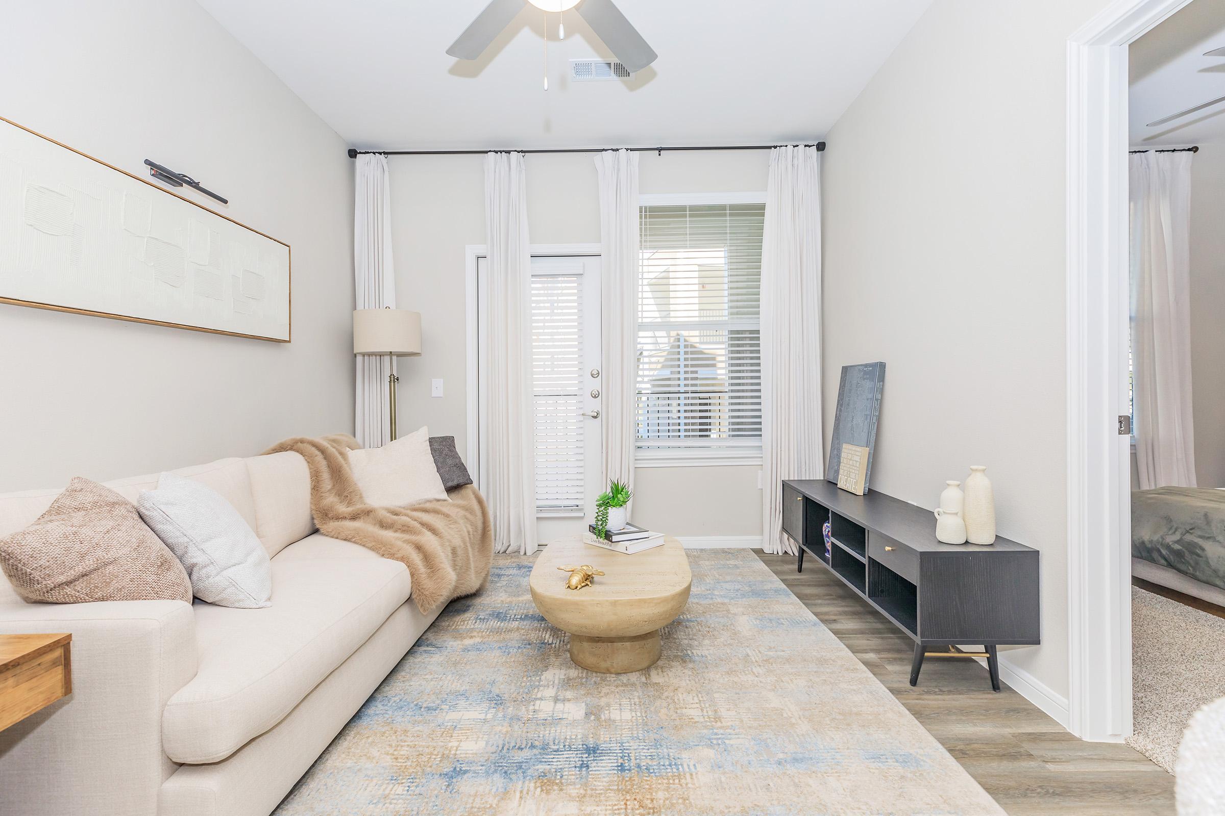 A cozy living room featuring a light-colored sofa with a throw blanket, a round coffee table, and a minimalist sideboard. Natural light filters through sheer curtains, and a door leads to an outdoor area. The decor includes subtle art on the wall and decorative items on the furniture, creating a welcoming atmosphere.
