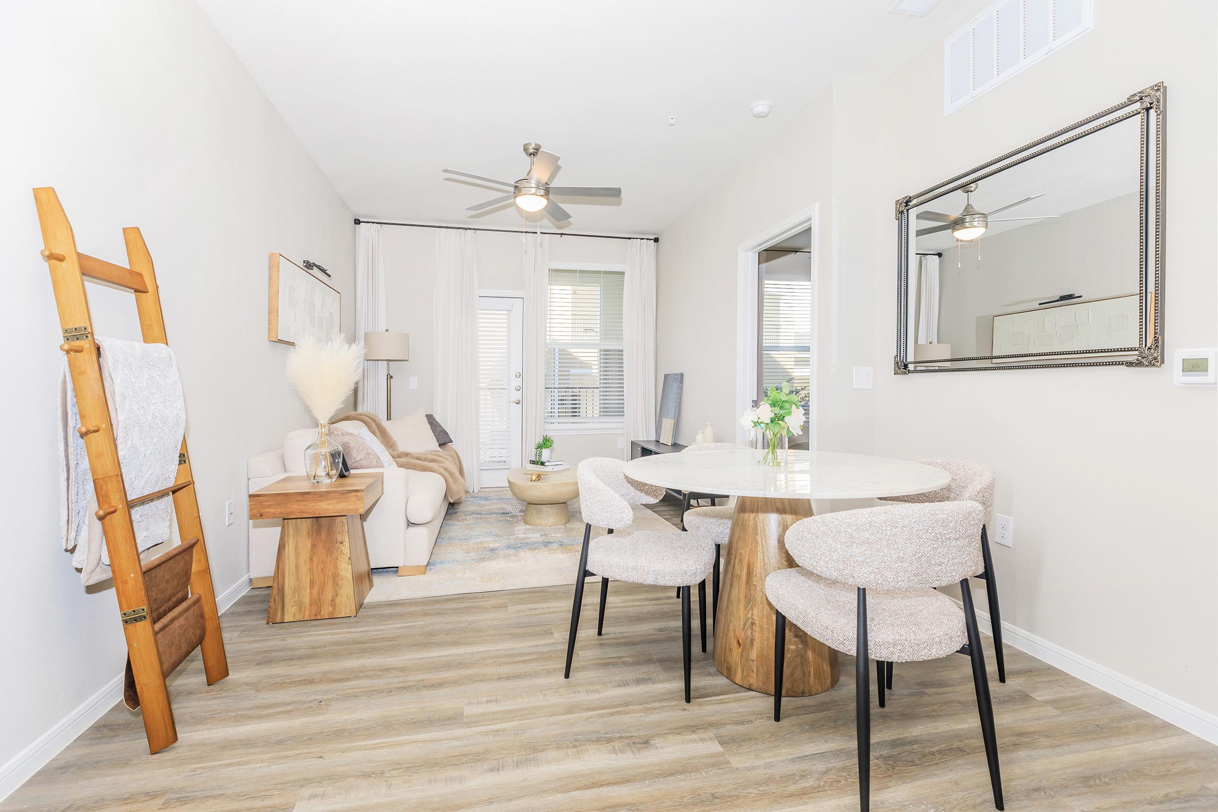 A modern living space featuring a cozy seating area with a white sofa and decorative pillows, a circular dining table with four upholstered chairs, and a stylish ladder shelf. Natural light enters through sliding glass doors, enhancing the warm, inviting atmosphere with wooden accents and neutral tones.