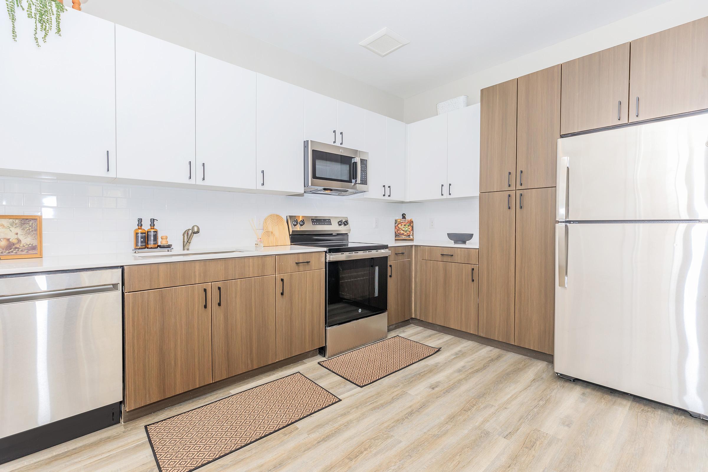Modern kitchen featuring a mix of white and wooden cabinets, stainless steel appliances including a microwave and oven, and a dishwasher. The countertops are clean with minimal decor, and there are two area rugs on the floor. Natural light brightens the space, creating a welcoming atmosphere.