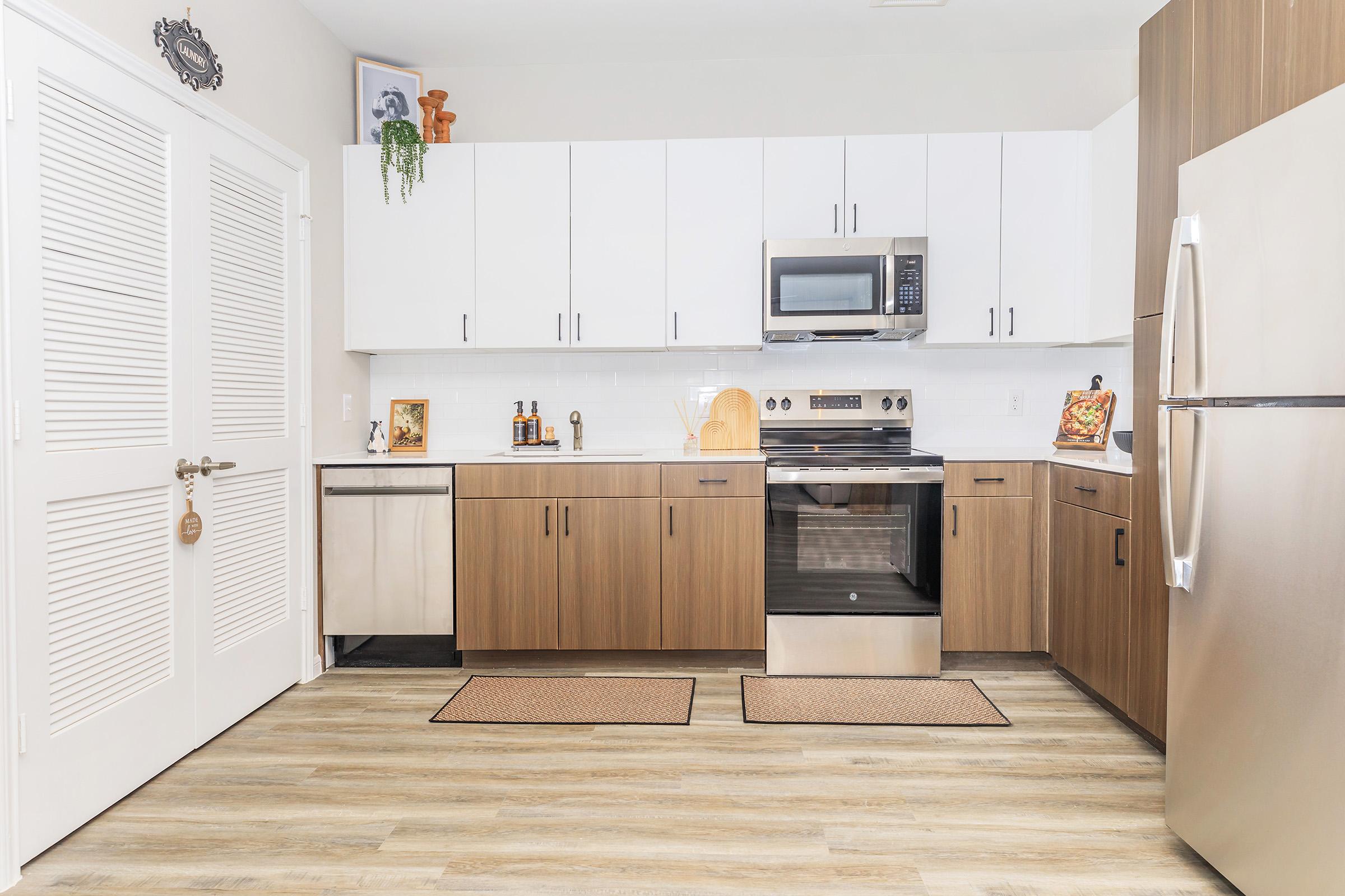 Modern kitchen featuring a mix of white upper cabinets and brown lower cabinets, stainless steel appliances including a refrigerator and microwave, and a smooth top stove. There are two textured mats on the floor and decorative elements on the countertop, enhancing the contemporary design.