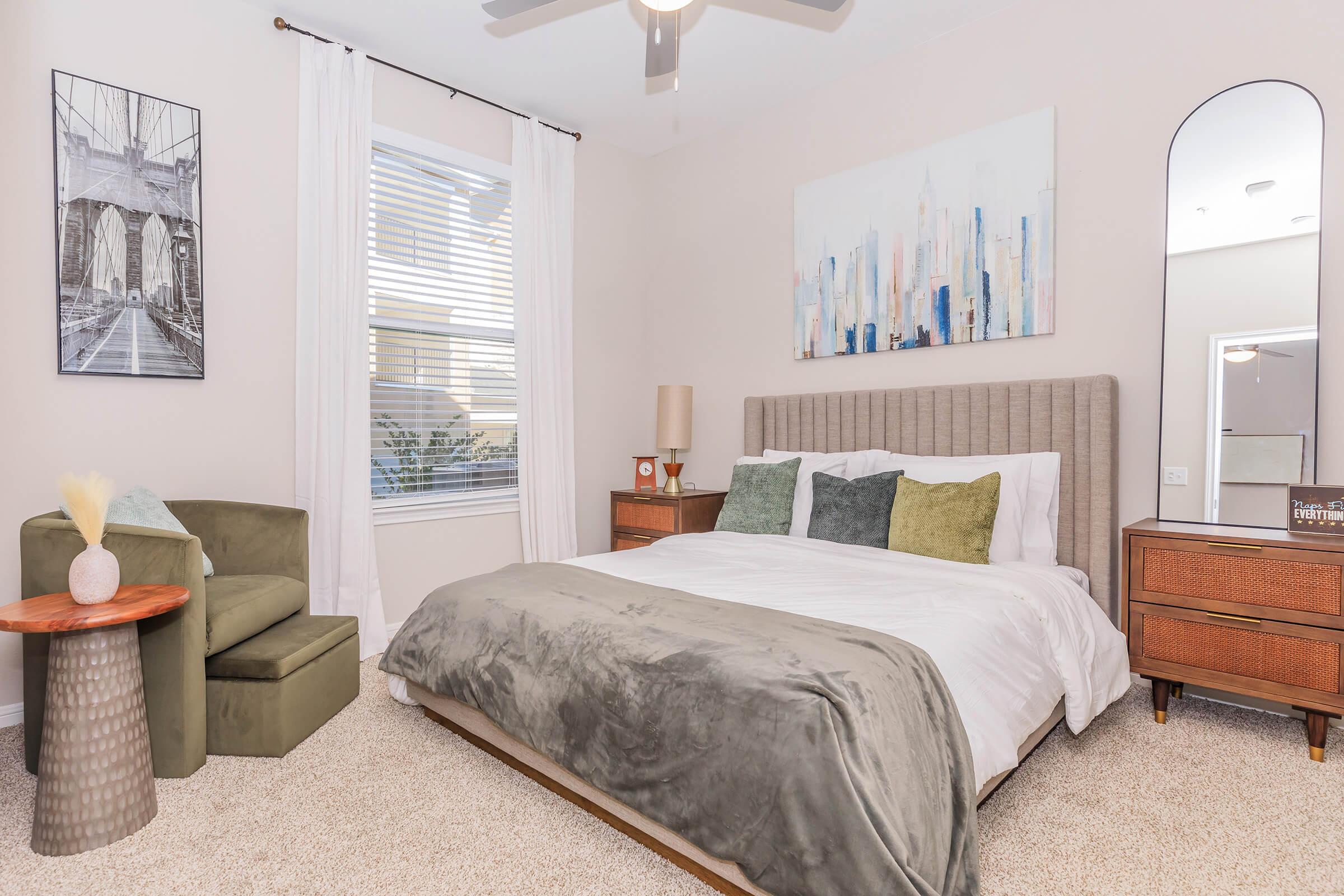 A cozy bedroom featuring a large bed with a grey comforter and decorative pillows. There is a green accent chair and a round side table with decorative elements. The decor includes a cityscape painting and a tall mirror, with light filtering through window blinds. The carpet adds warmth to the space.