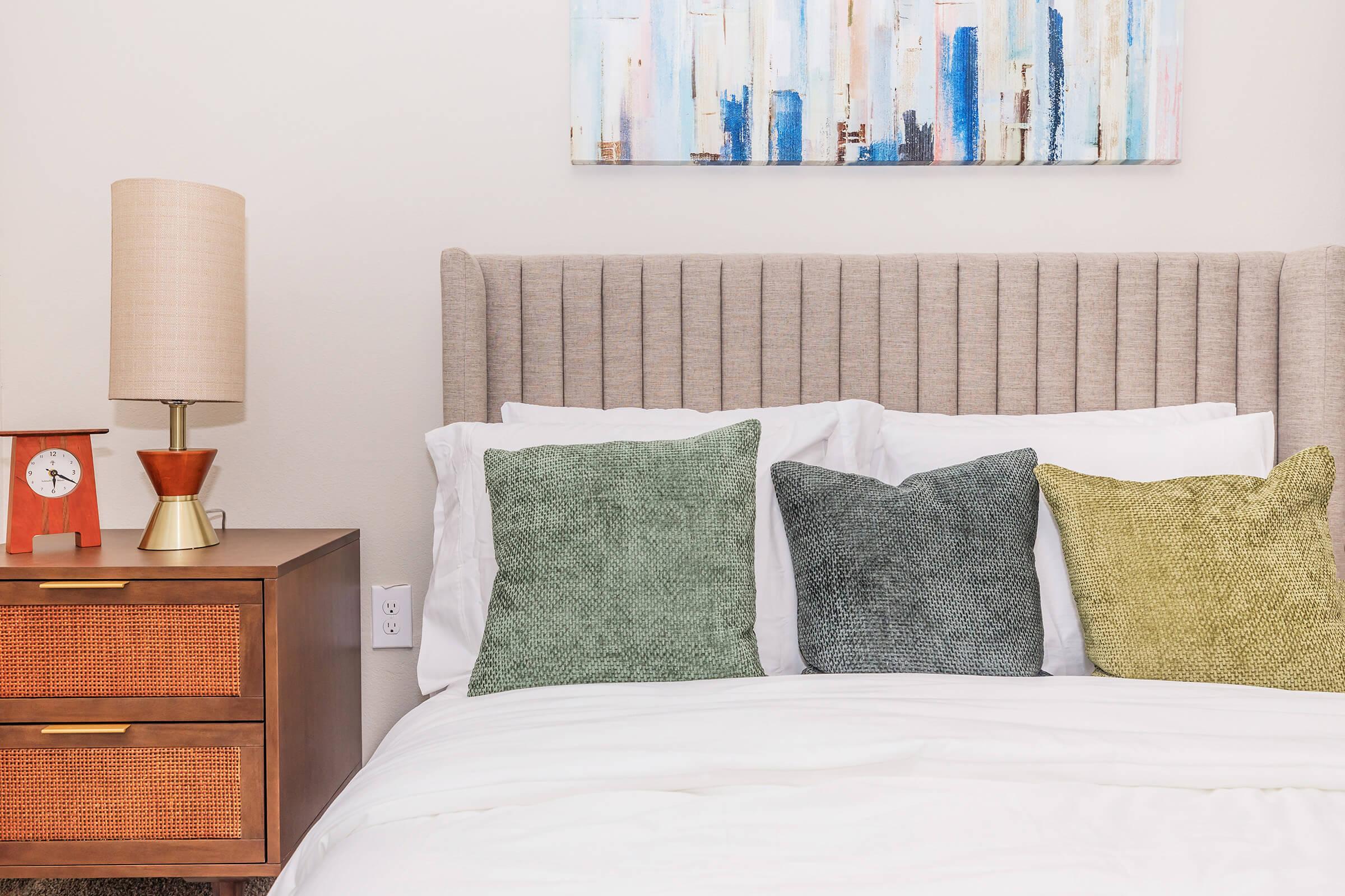 A neatly made bed with white linens and three decorative pillows in shades of green and yellow. A stylish nightstand with a clock and a lamp featuring a textured lampshade are positioned beside the bed. An abstract painting in blue and beige tones hangs on the wall above.