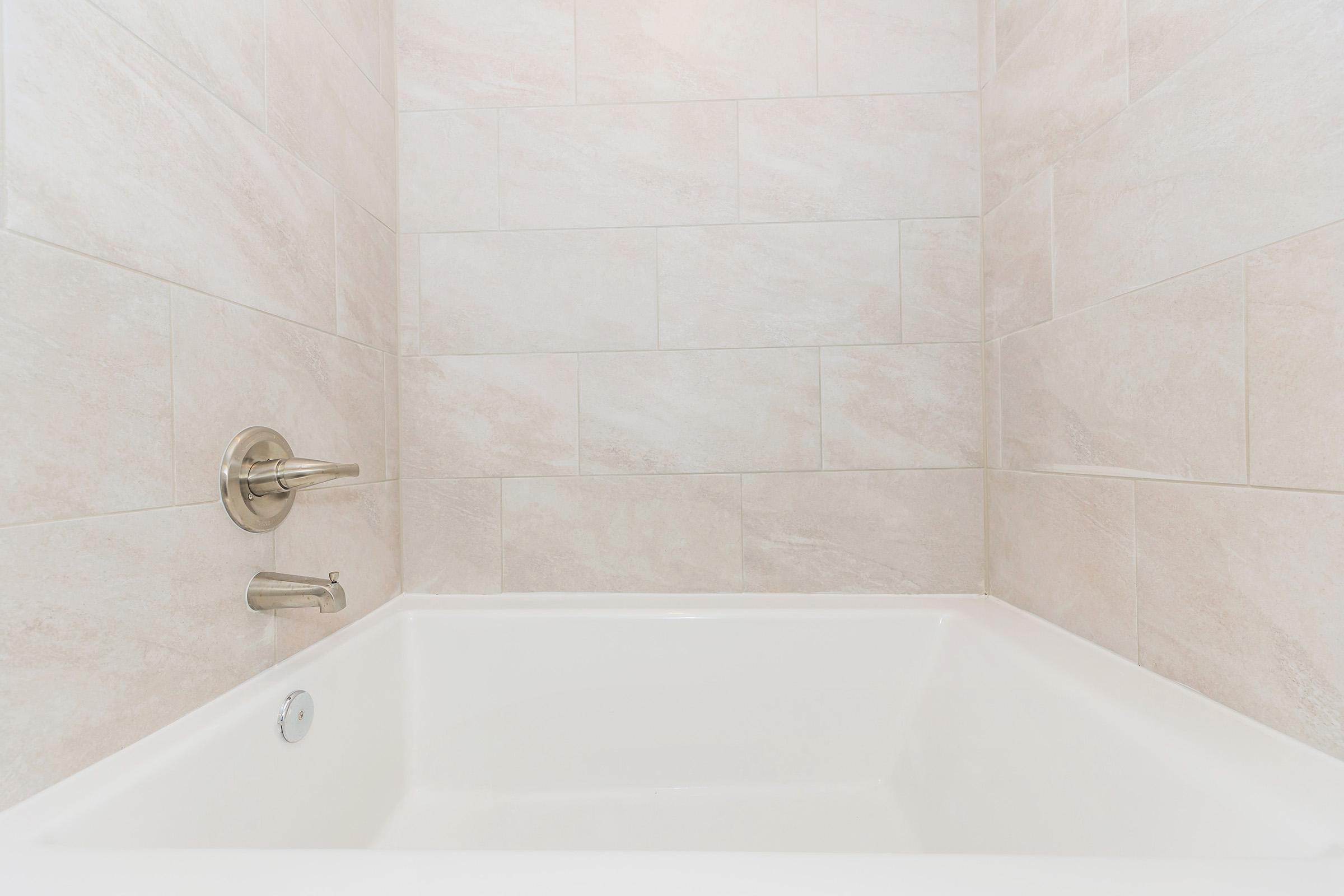 A clean, modern bathtub set against light-colored, textured tile walls. The tub is white, with a silver faucet and control handle, and a plain, spacious design, suitable for a minimalist bathroom aesthetic. The overall appearance is bright and inviting.