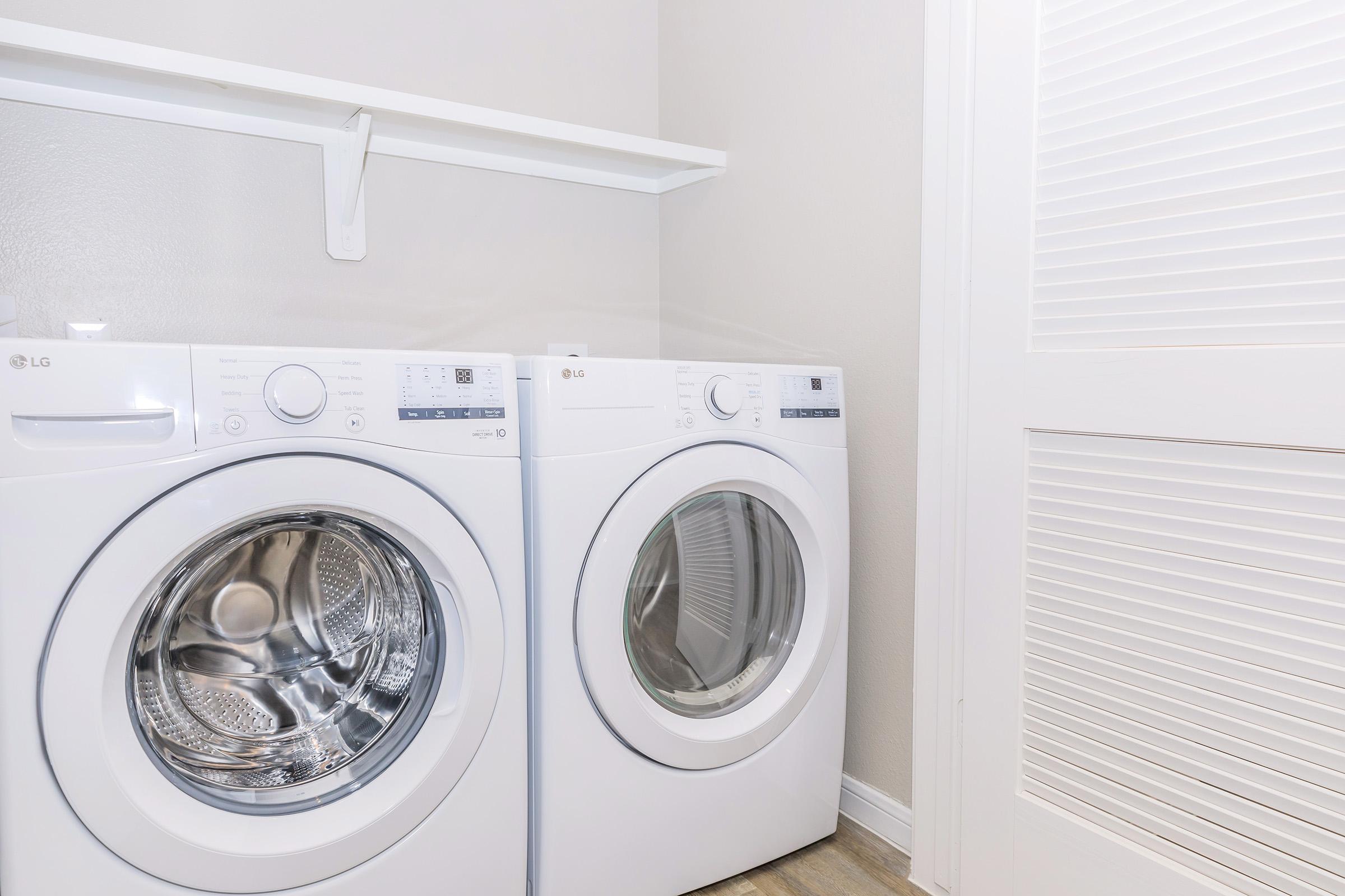Two modern white washing machines, one front-loading and one top-loading, positioned side by side in a clean laundry room. The walls are light-colored, and there is a white shelf above the machines. A door with slats is visible in the background, adding to the tidy appearance of the space.