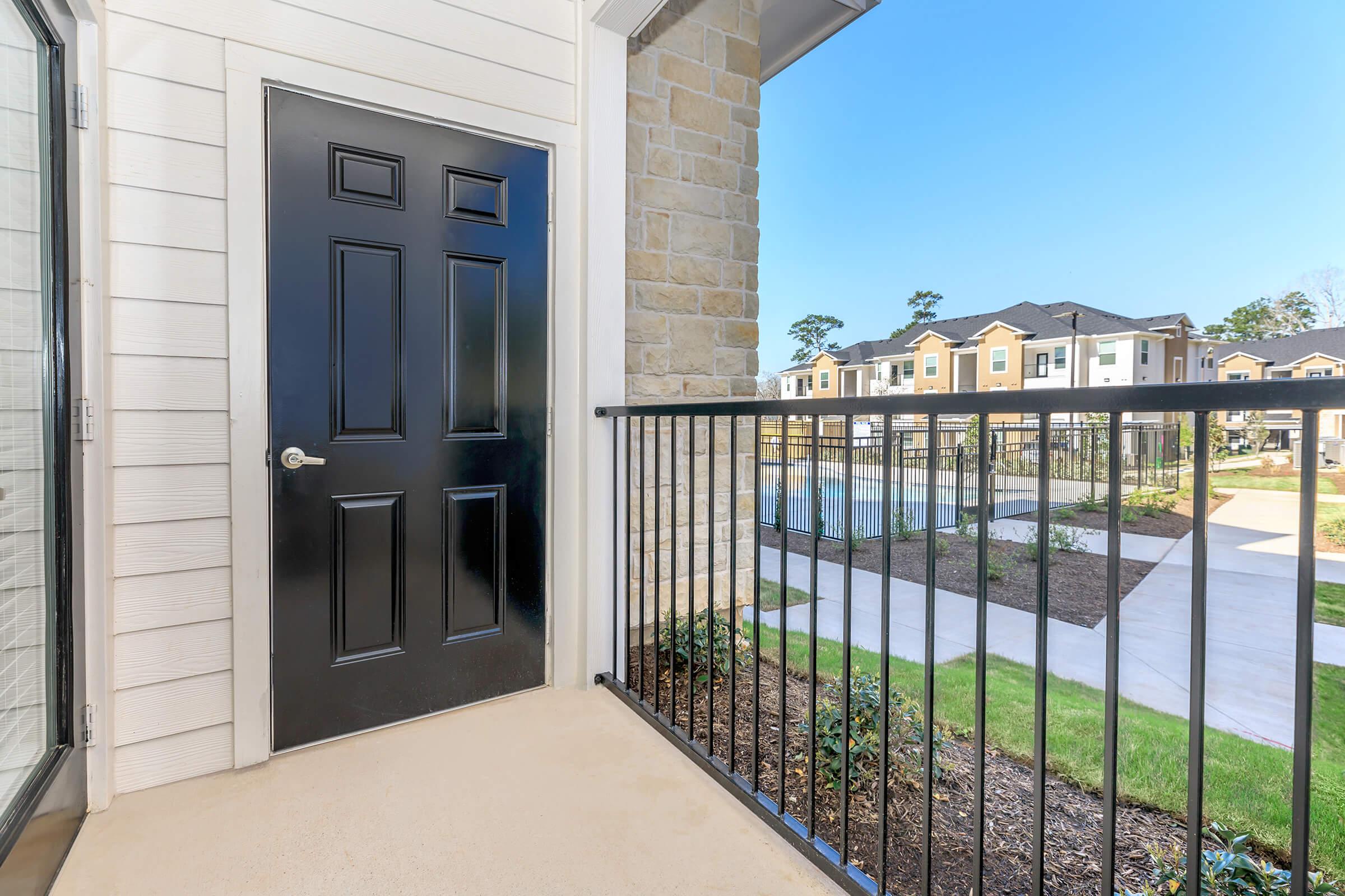 A black door on a balcony with a metal railing, overlooking a landscaped area of an apartment complex. The view features green grass, pathways, and multiple residential buildings in the background under a clear blue sky.