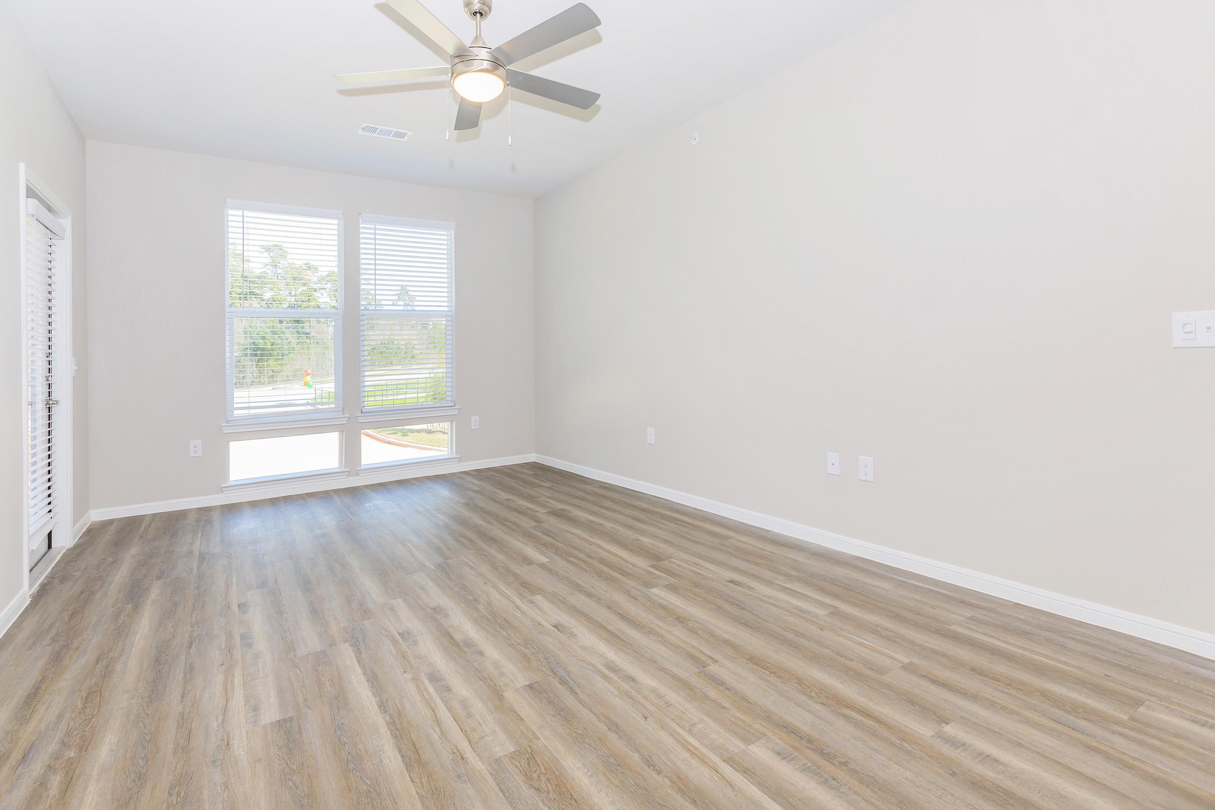 Spacious interior of a room with hardwood-style flooring, a ceiling fan, and large windows allowing natural light. The walls are painted a light color, creating an airy atmosphere. The room is empty, suggesting it is ready for furnishing or customization.